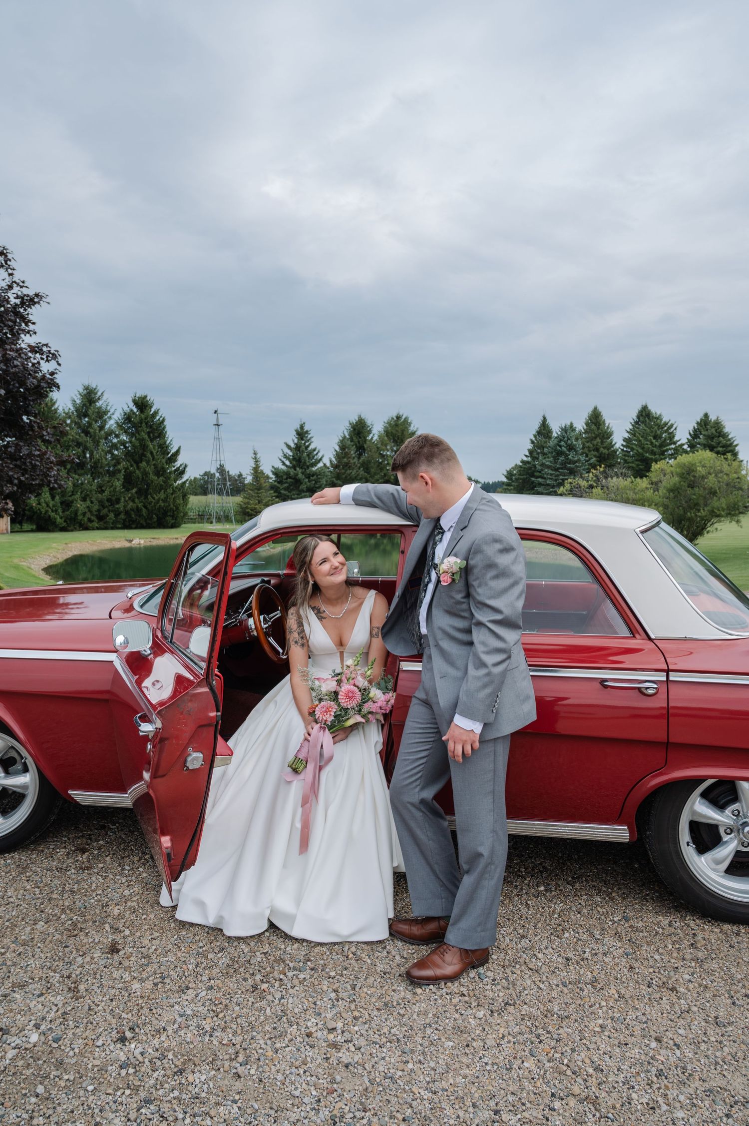 Bride and groom pose beside a classic red vintage car on their wedding day, surrounded by lush green trees.