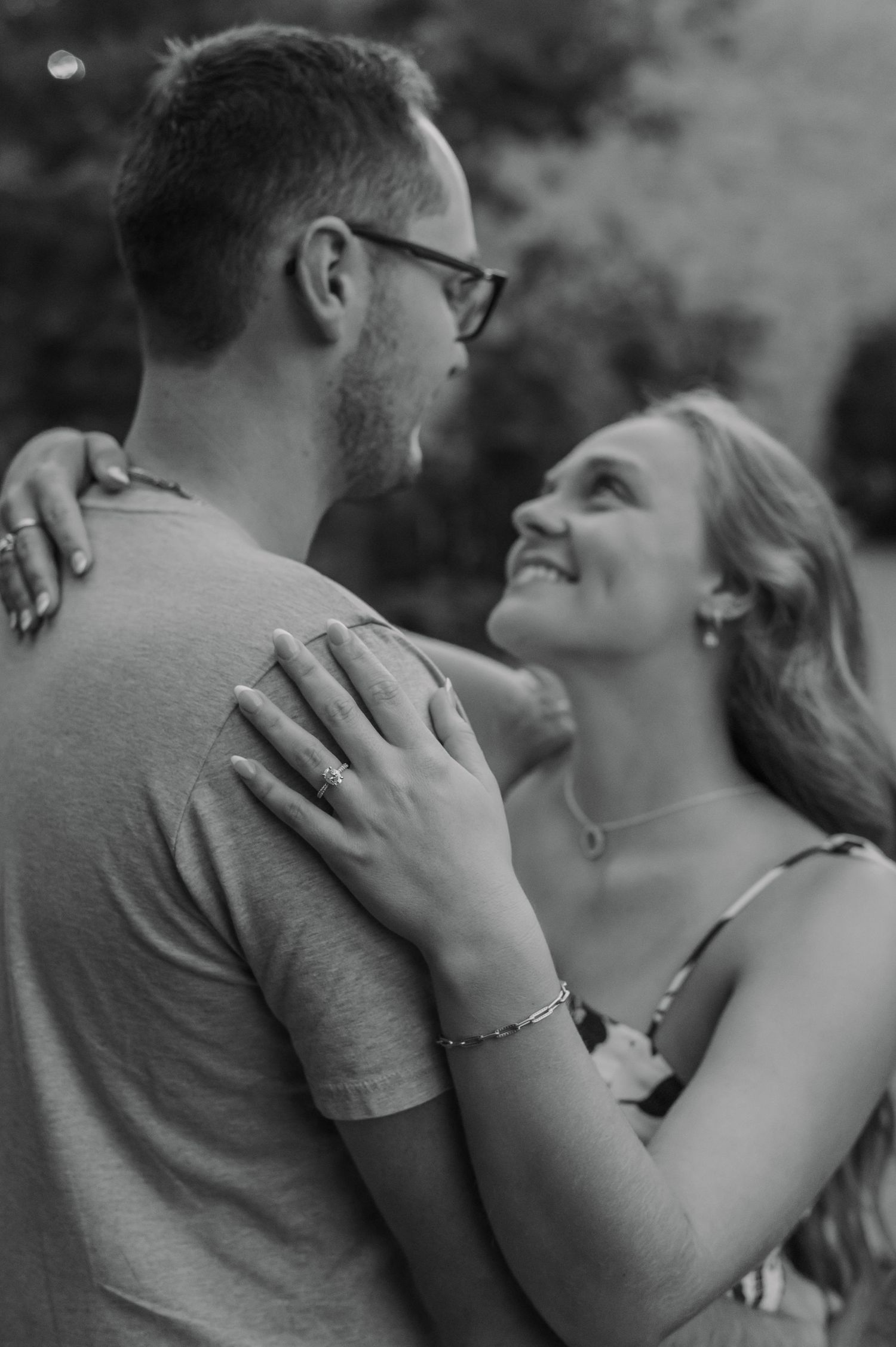 Black and white photo of a smiling couple embracing outdoors, woman wearing bracelet and ring looks lovingly at man.