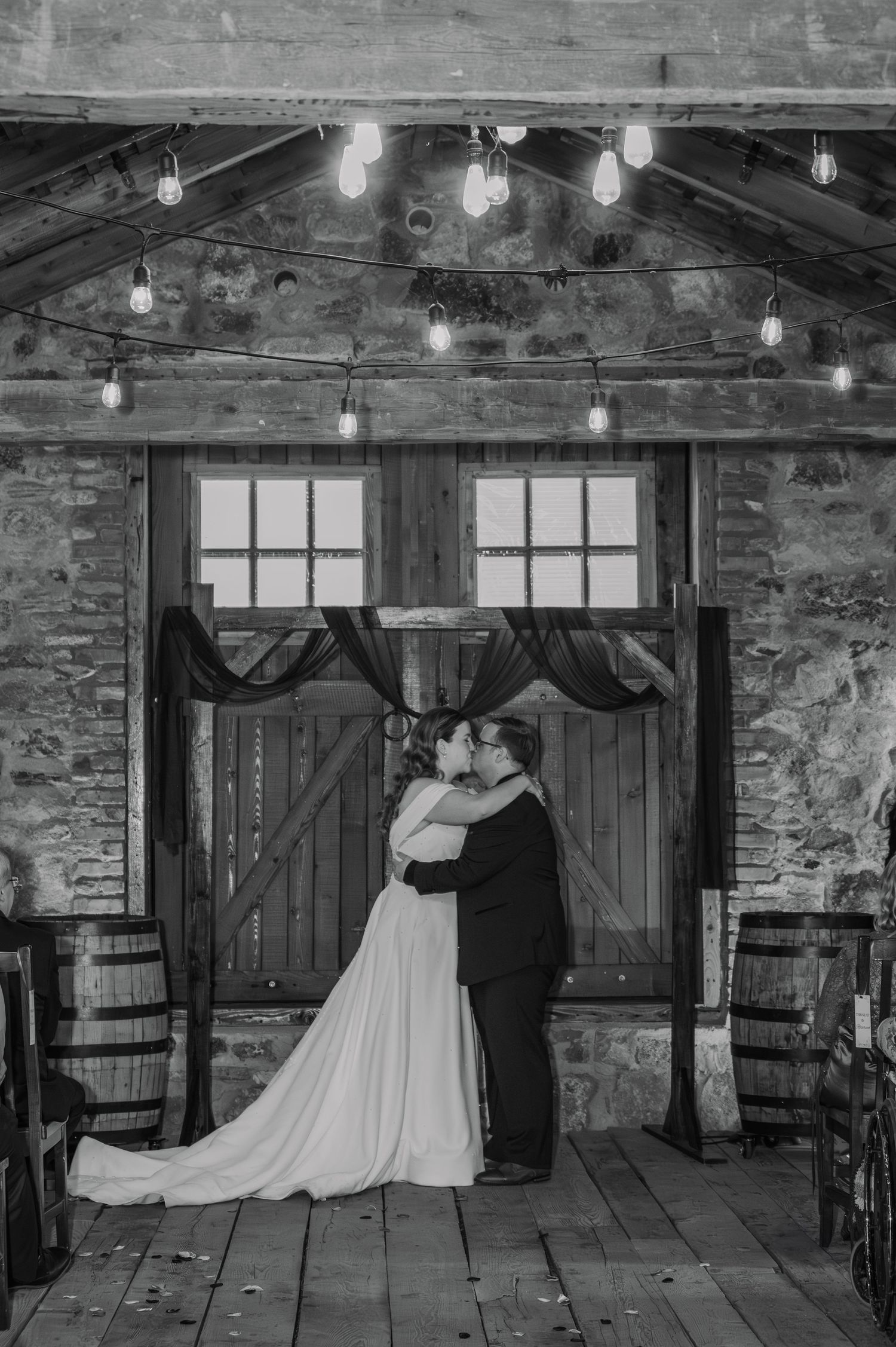 Black and white photo of a bride and groom sharing a kiss in a rustic barn venue with string lights overhead.
