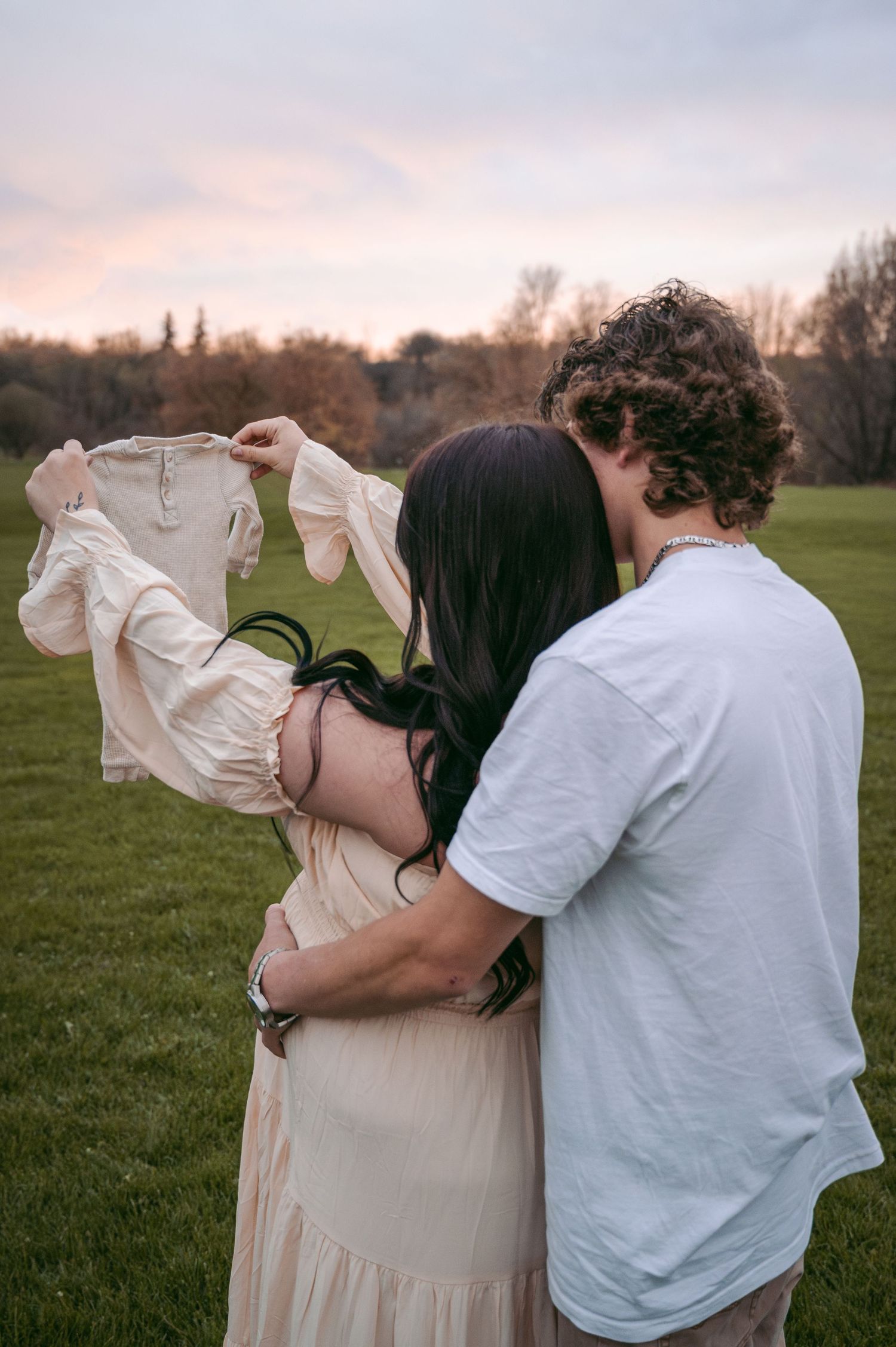Couple holding tiny baby outfit during outdoor maternity photography session.