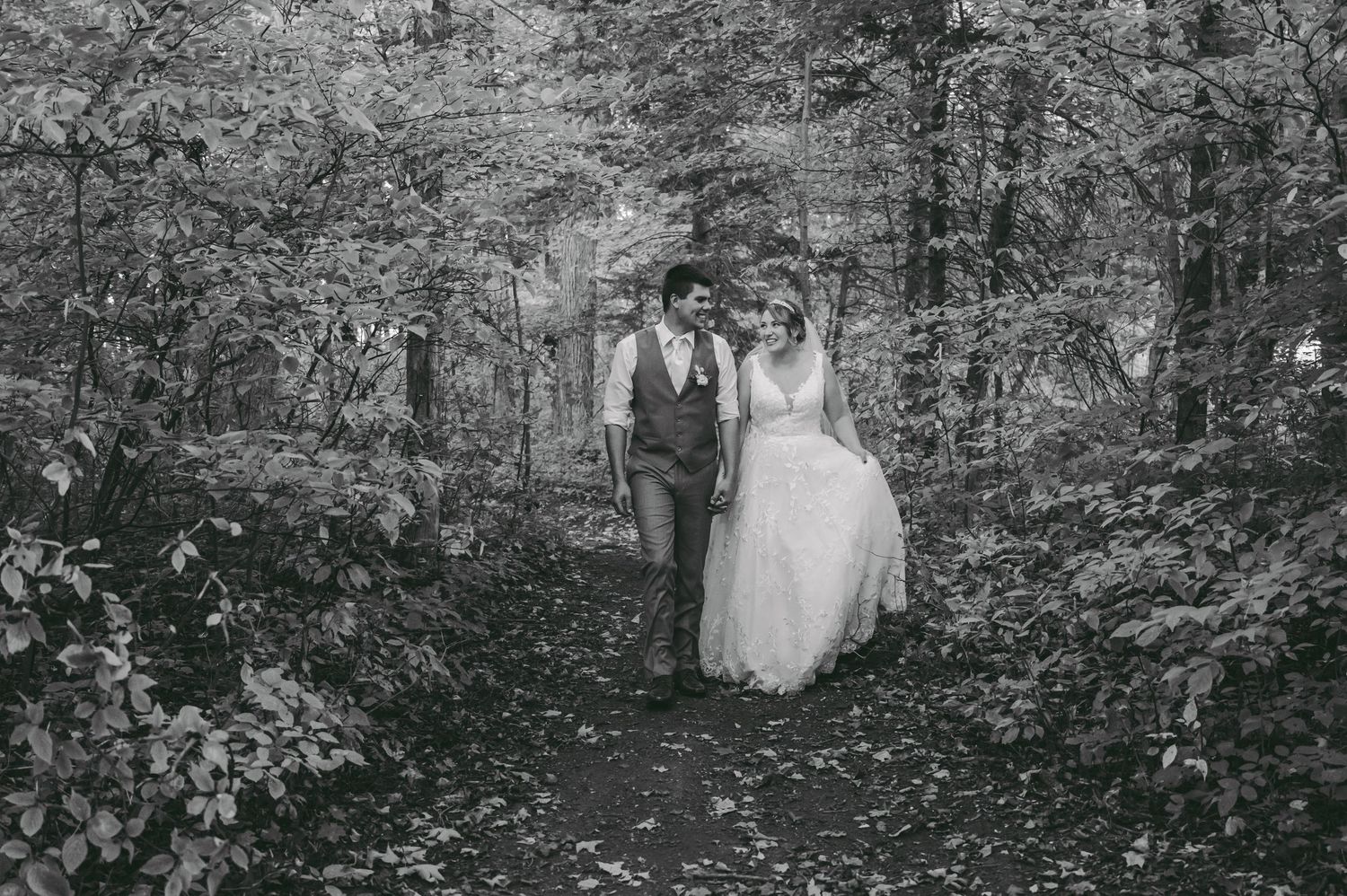 Black and white photo of couple walking hand in hand through wooded forest path in wedding attire.