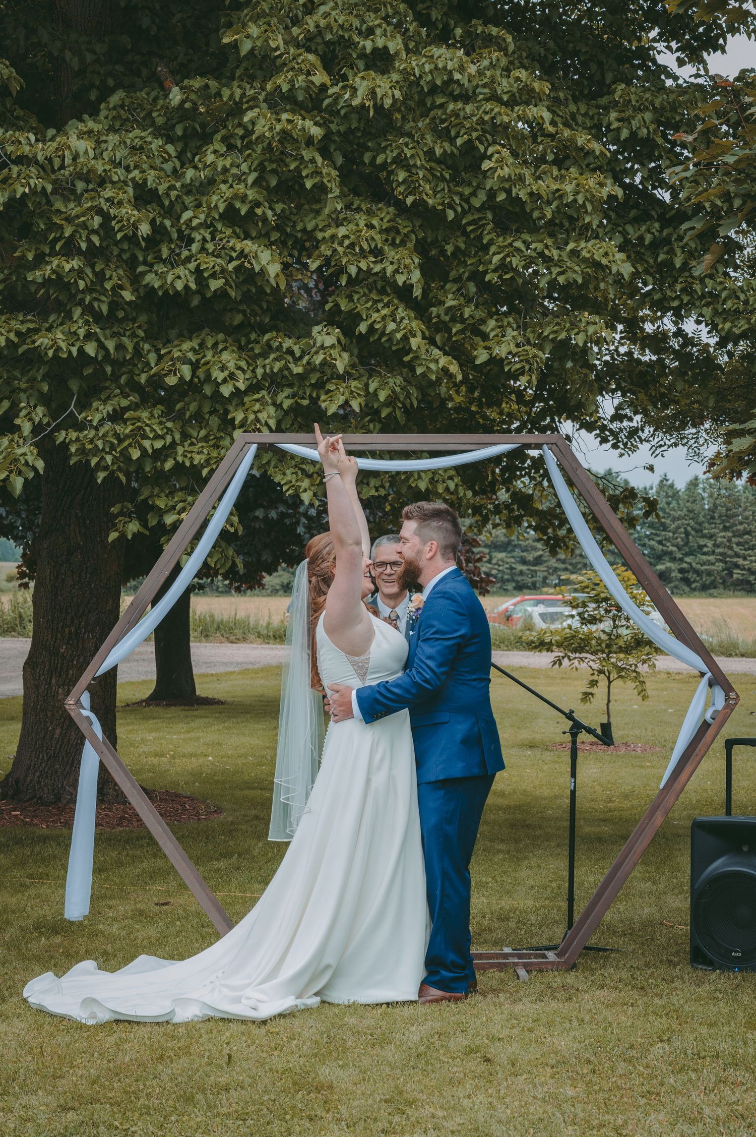 Newlyweds celebrate under geometric wooden arch at outdoor ceremony.