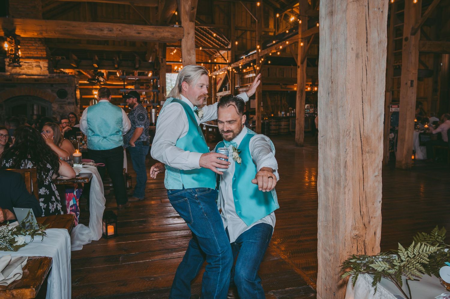 Wedding guests dancing and celebrating at a rustic barn reception with wooden beams and string lights.
