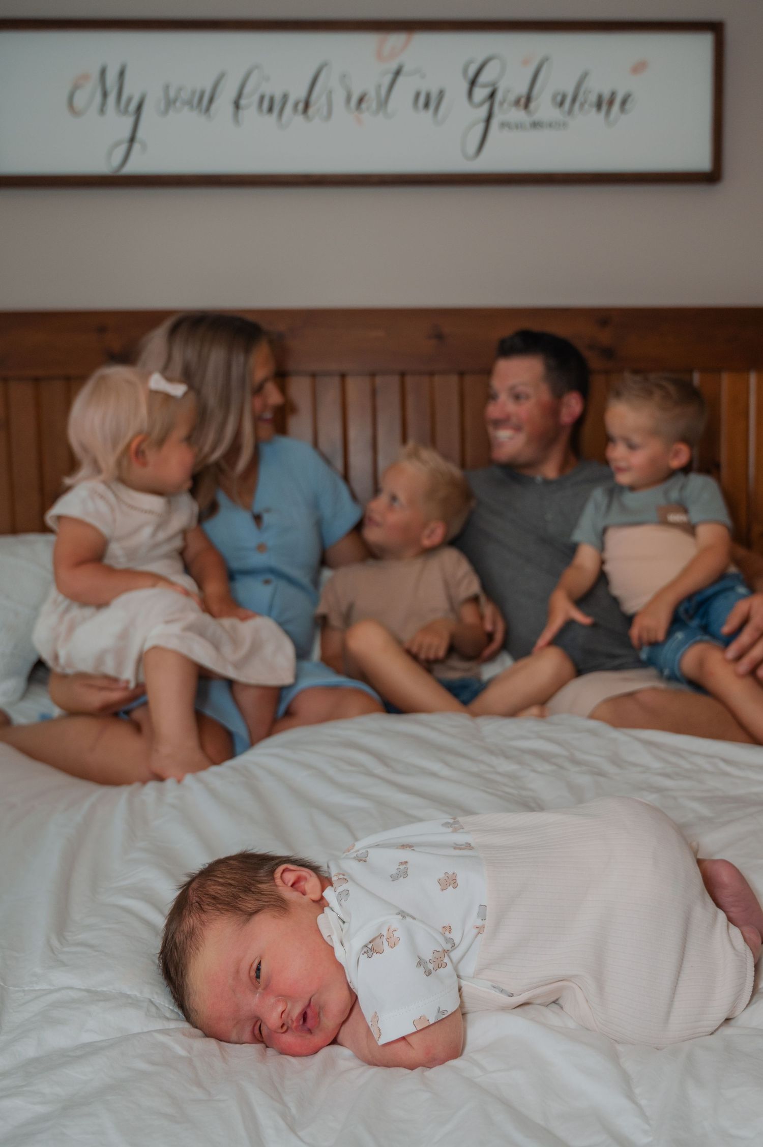 A sleeping baby in white sleepwear resting peacefully on white bedding, with their family in the background.