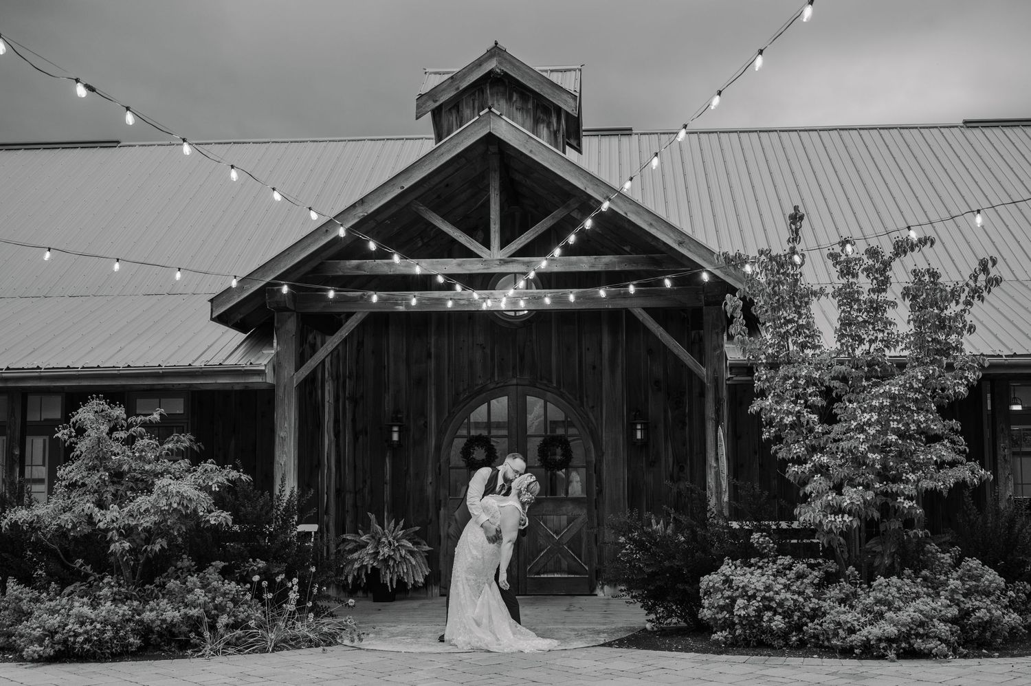 Black and white photo of a wedding couple embracing outside a rustic barn venue adorned with string lights.