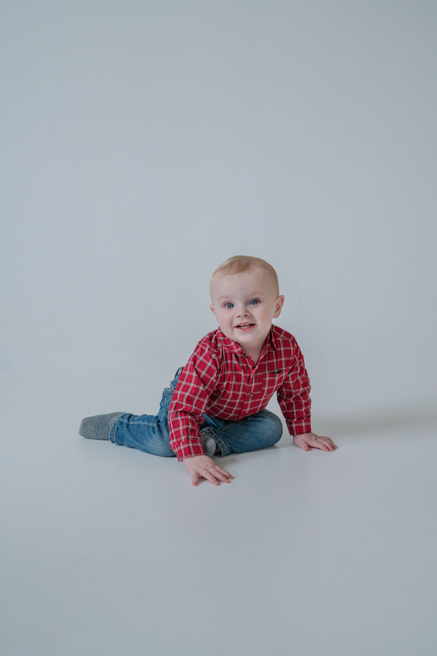 A cheerful baby boy in a red plaid shirt and jeans sits on a white studio floor, looking up with a bright smile.