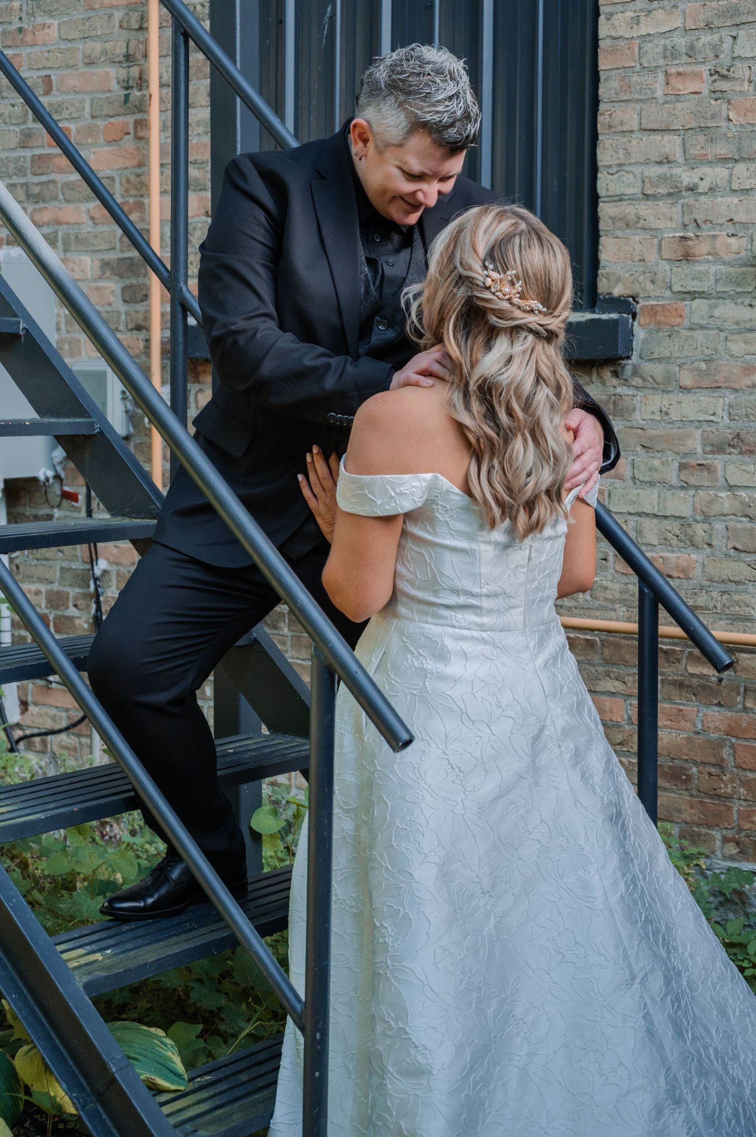 A couple in wedding attire share an intimate moment on an outdoor staircase with brick wall backdrop.
