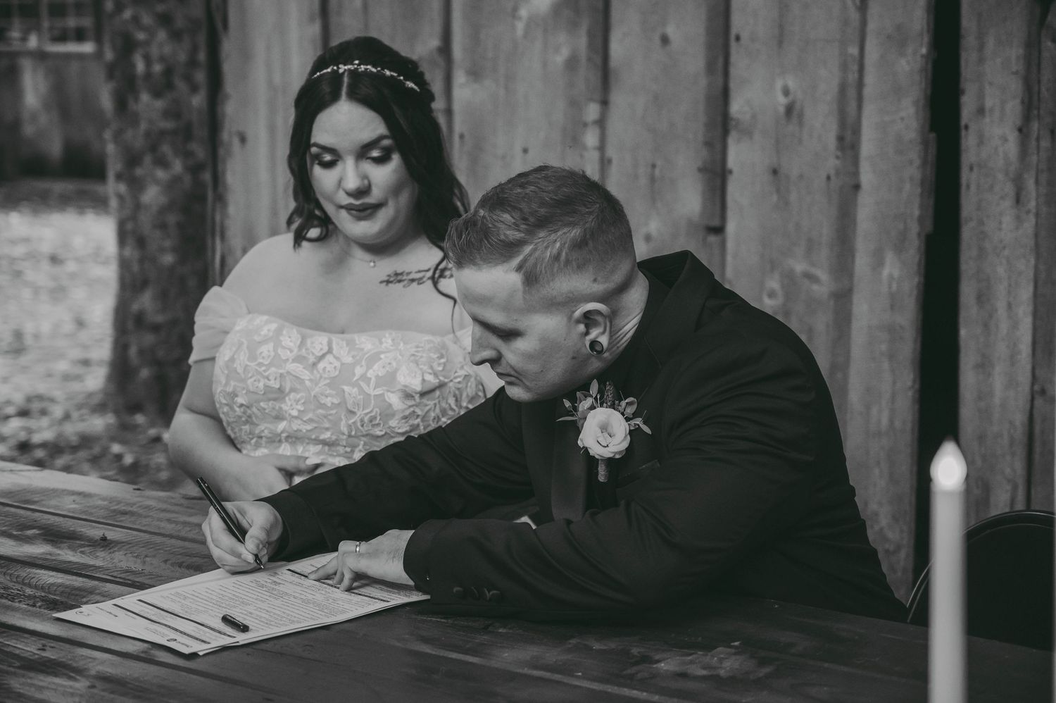 Bride and groom sign wedding documents together at a wooden table in black and white.