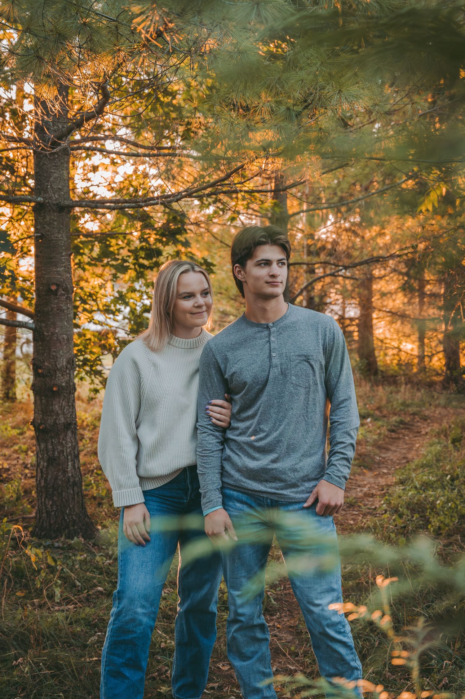 A couple poses together in a forest setting with warm autumn colors in the background.