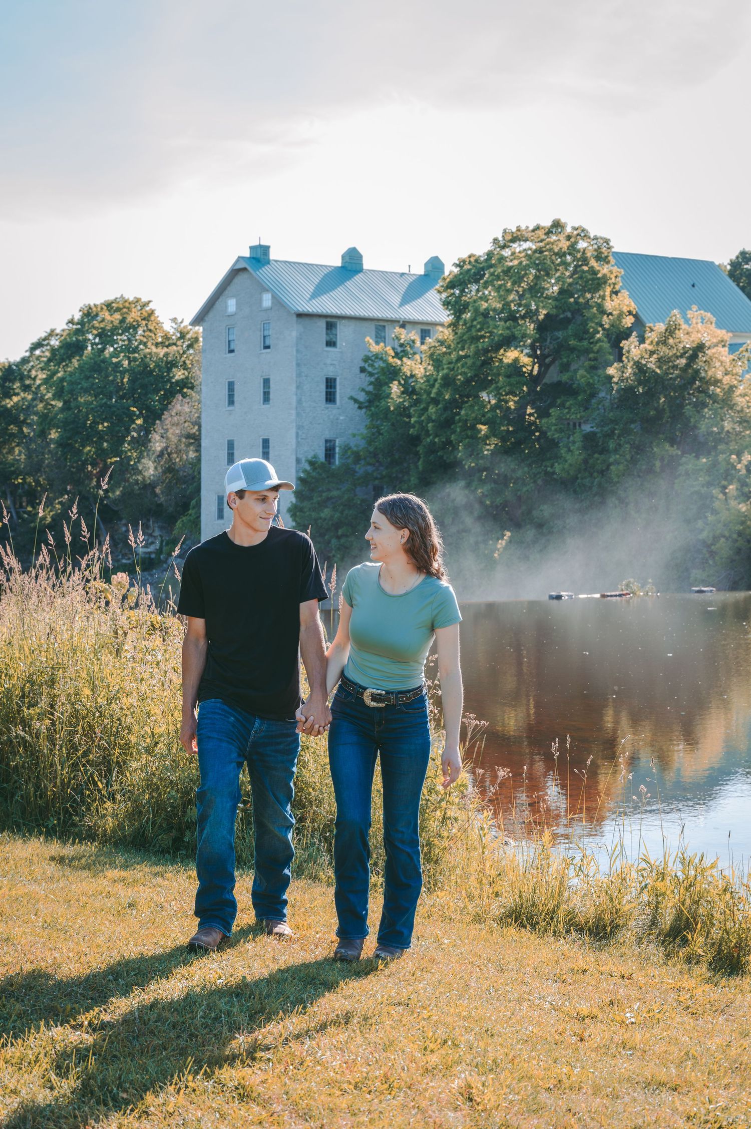 A couple walks hand in hand along a riverside path with a historic building in the background.
