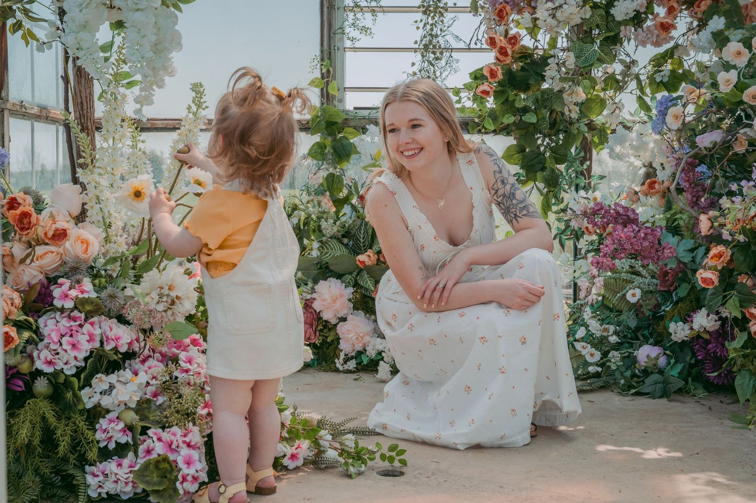 Playful interaction between adult and toddler surrounded by lush floral greenhouse display.