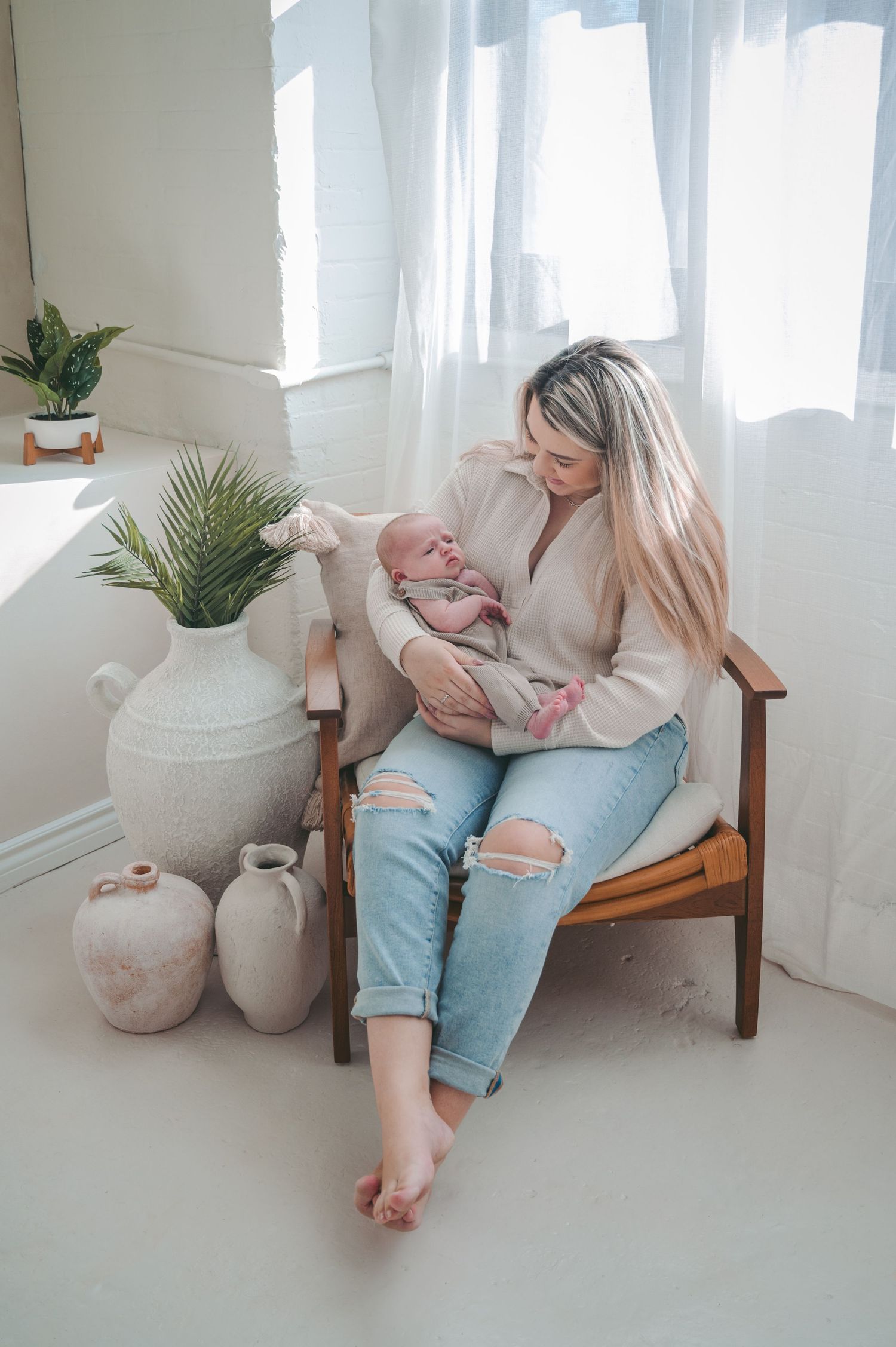 Parent holding baby in cozy corner with decorative vases and plants by window.