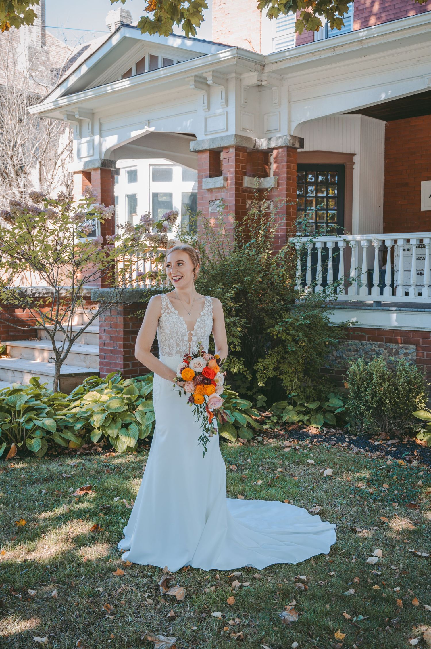 Bride in elegant white dress holds cascading orange bouquet in front of historic brick house.