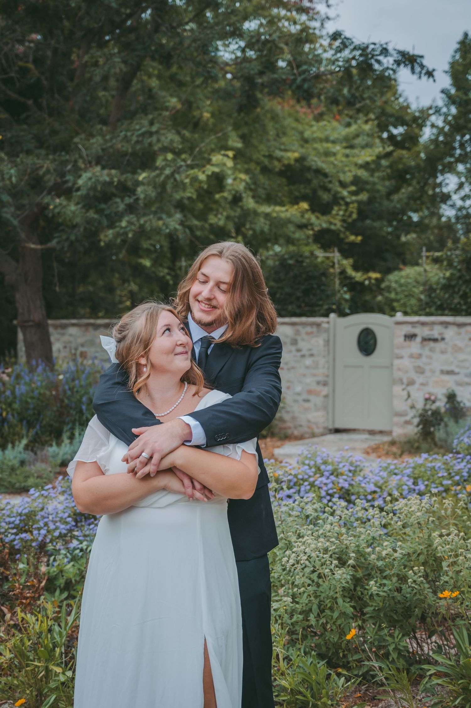 A groom in a navy suit embraces his bride in a white dress in a lush garden setting on their wedding day.