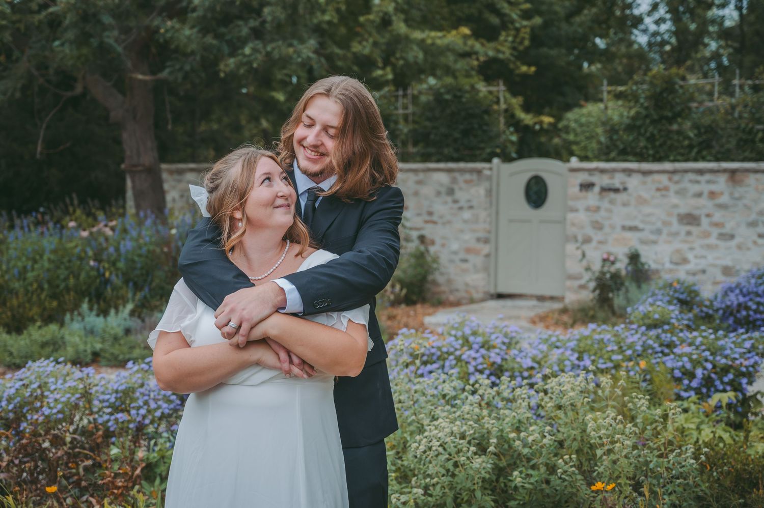 Couple embracing in a garden wedding setting with purple wildflowers and stone wall backdrop.