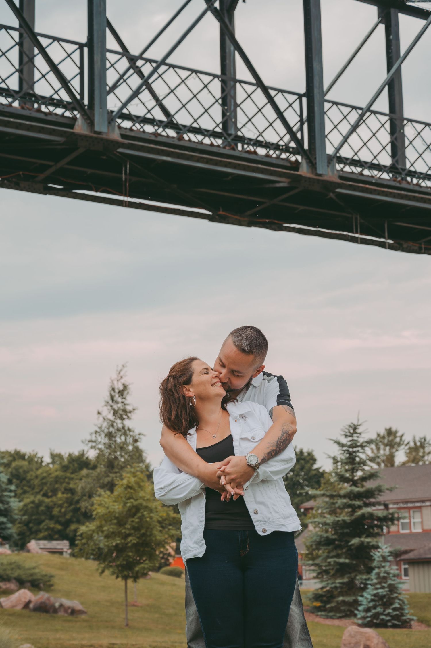 A romantic embrace under a steel bridge structure at sunset.