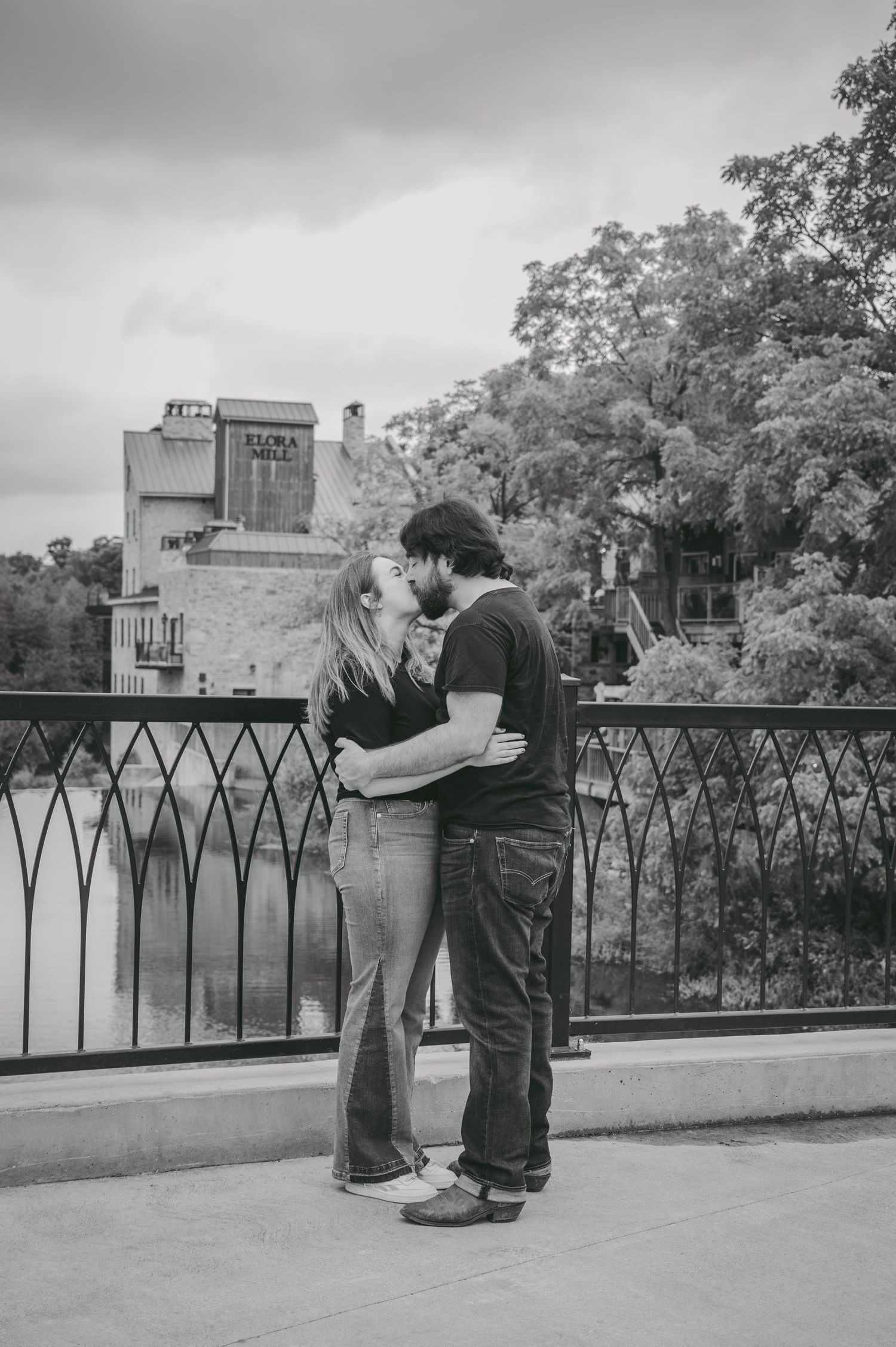 Black and white photo of couple embracing on urban balcony with historic Elora Mill building behind.