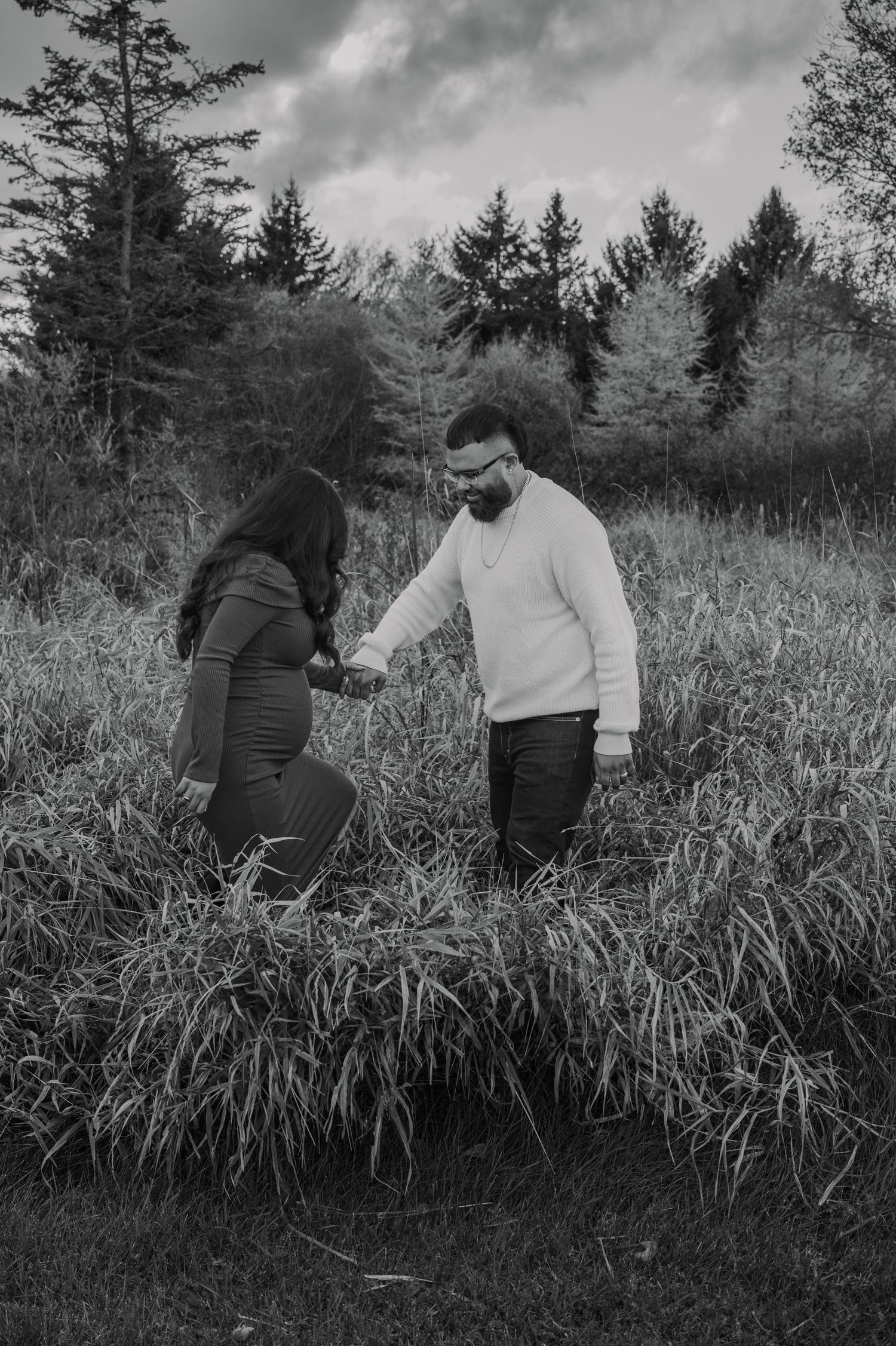 A couple in a field of tall grass share an intimate black and white moment against a backdrop of evergreen trees.