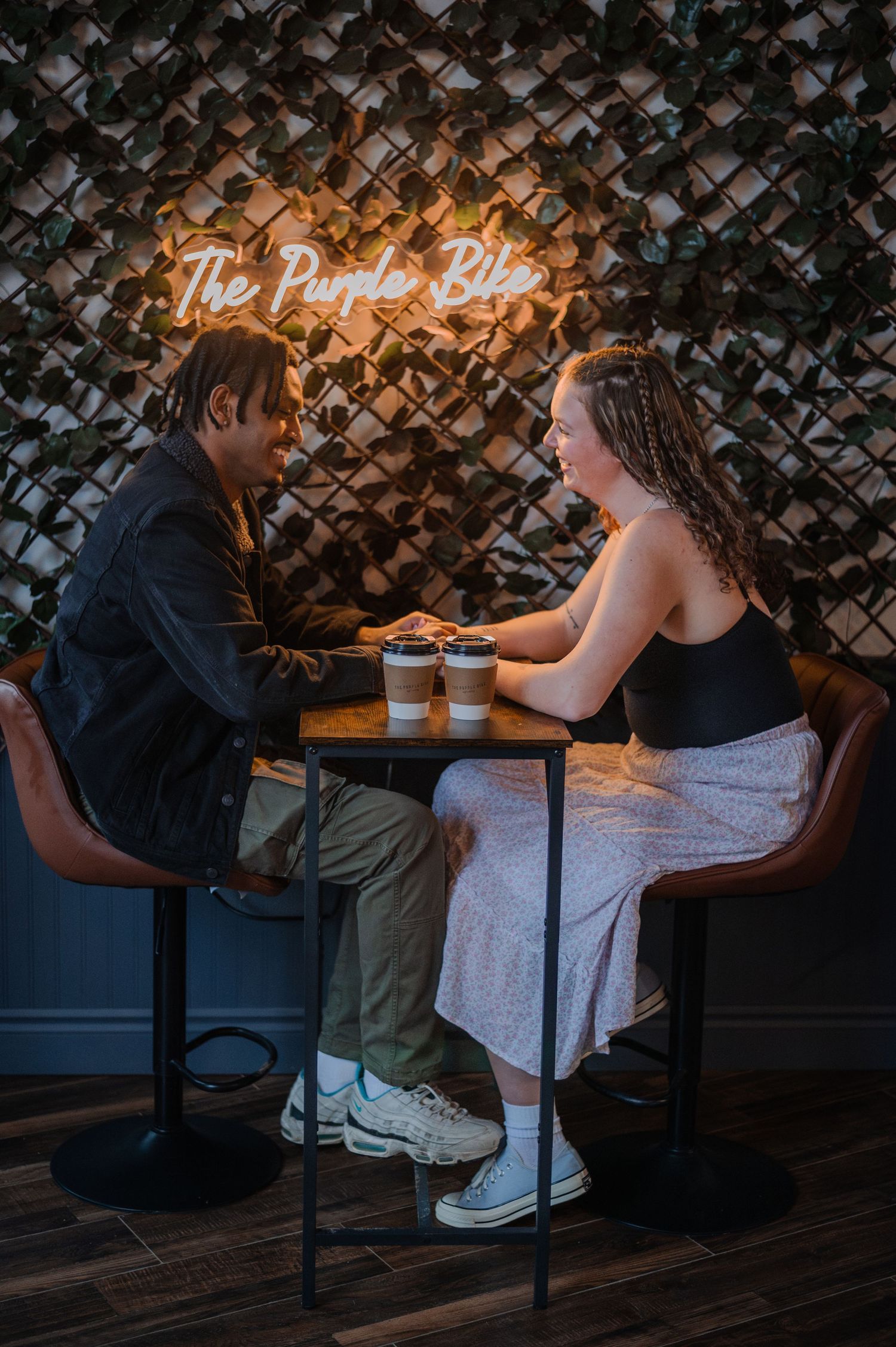 Two people share coffee at a small cafe table beneath a glowing neon sign reading The Purple Bike.