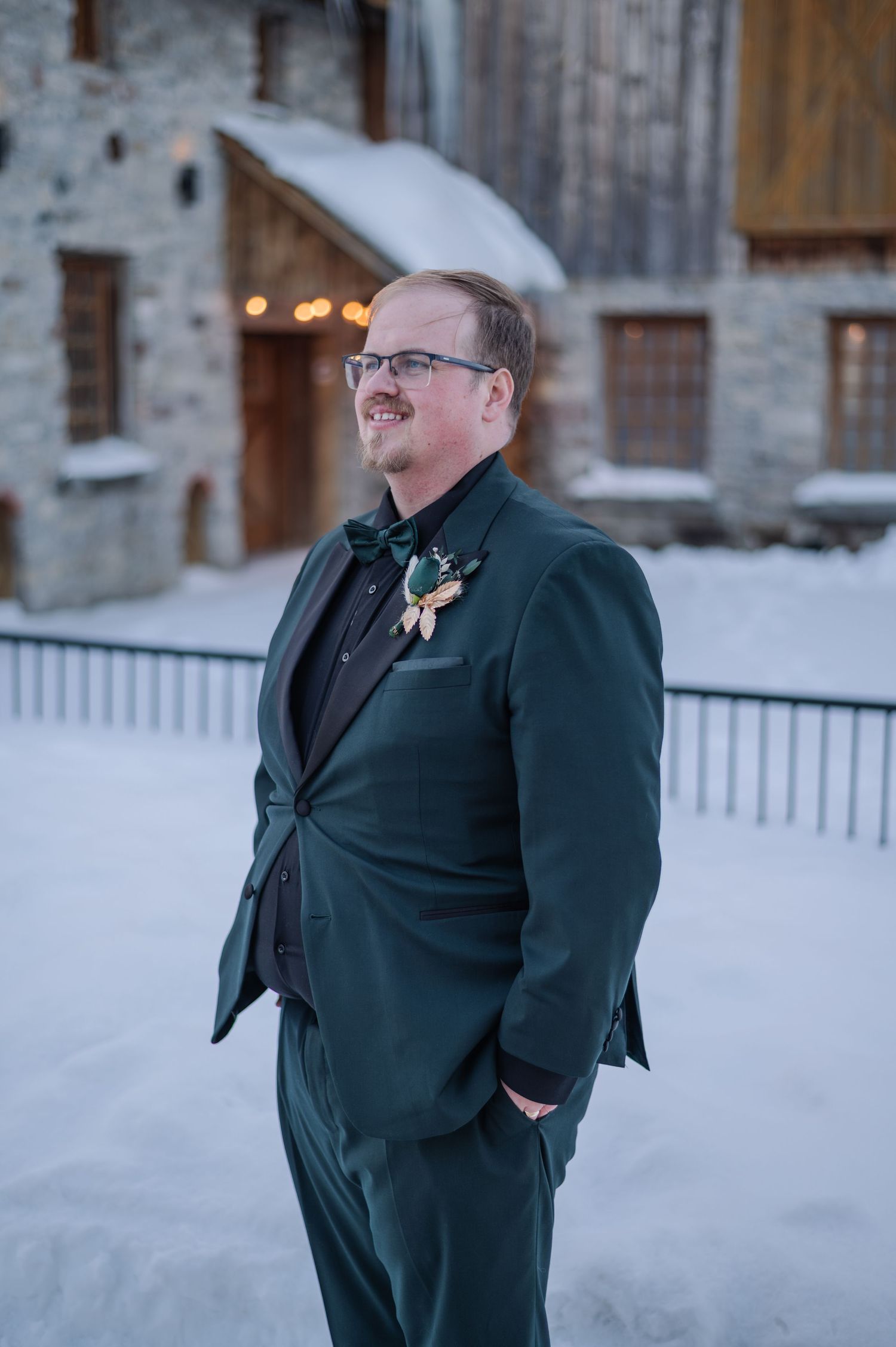 Person wearing a dark green suit and glasses stands in snowy winter setting near rustic Brussels Four Winds Barn.