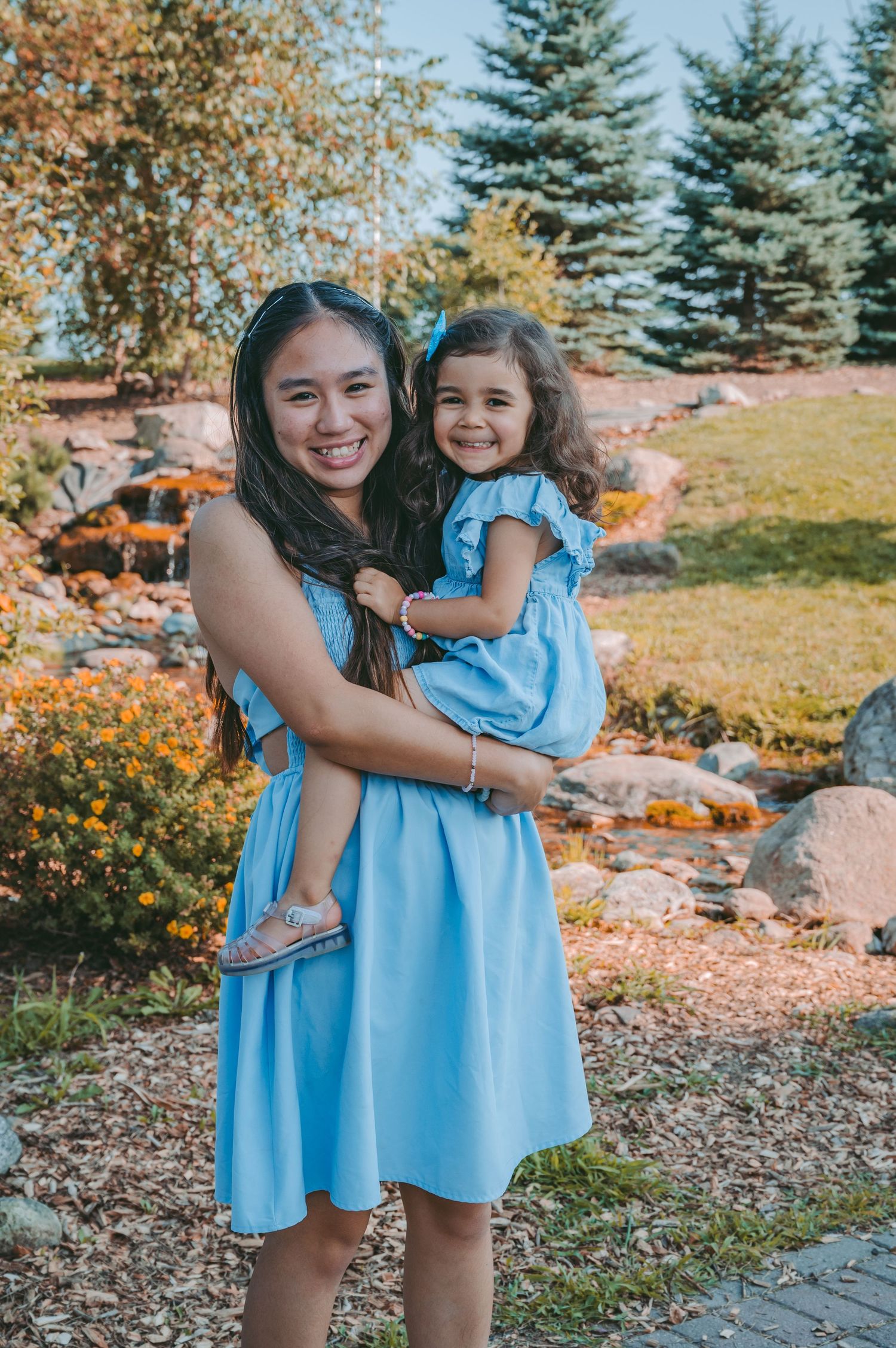 Siblings in matching blue dresses share a joyful embrace in a garden setting with evergreens.