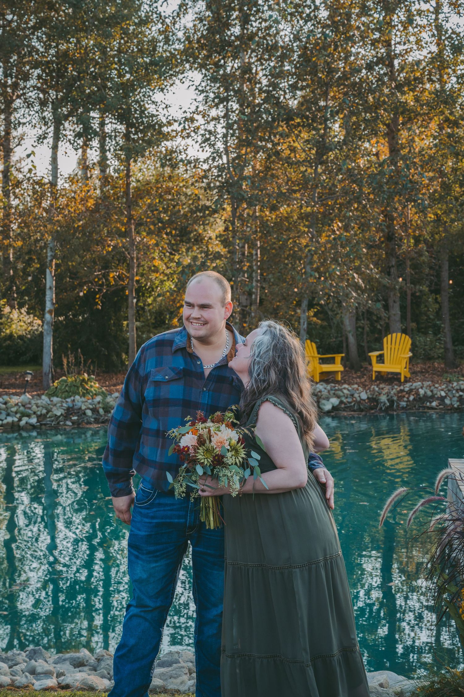 A couple poses by a turquoise pool surrounded by autumn trees with yellow chairs in the background.