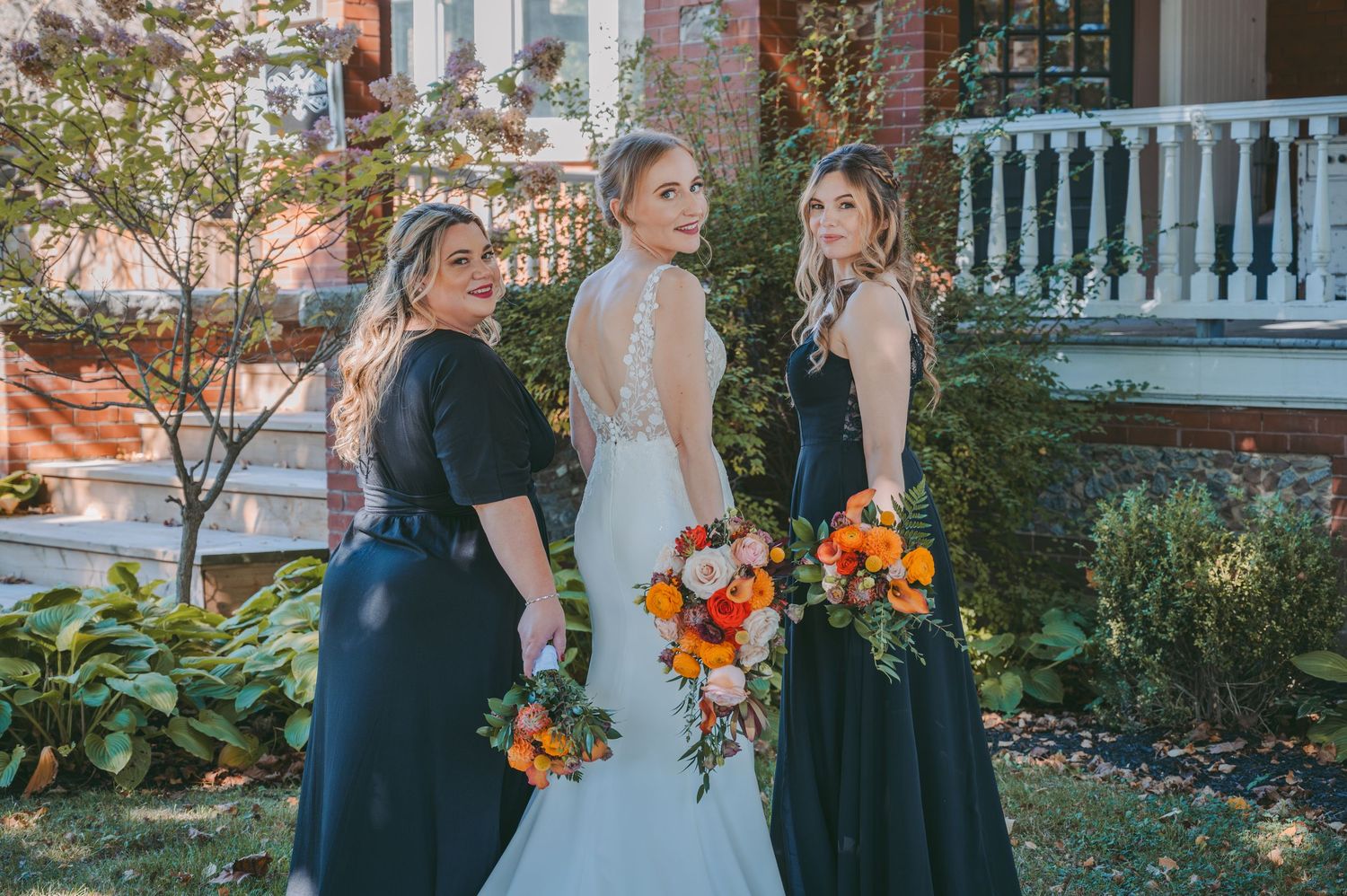 Three women hold orange and white bouquets while posing in front of a historic house porch.