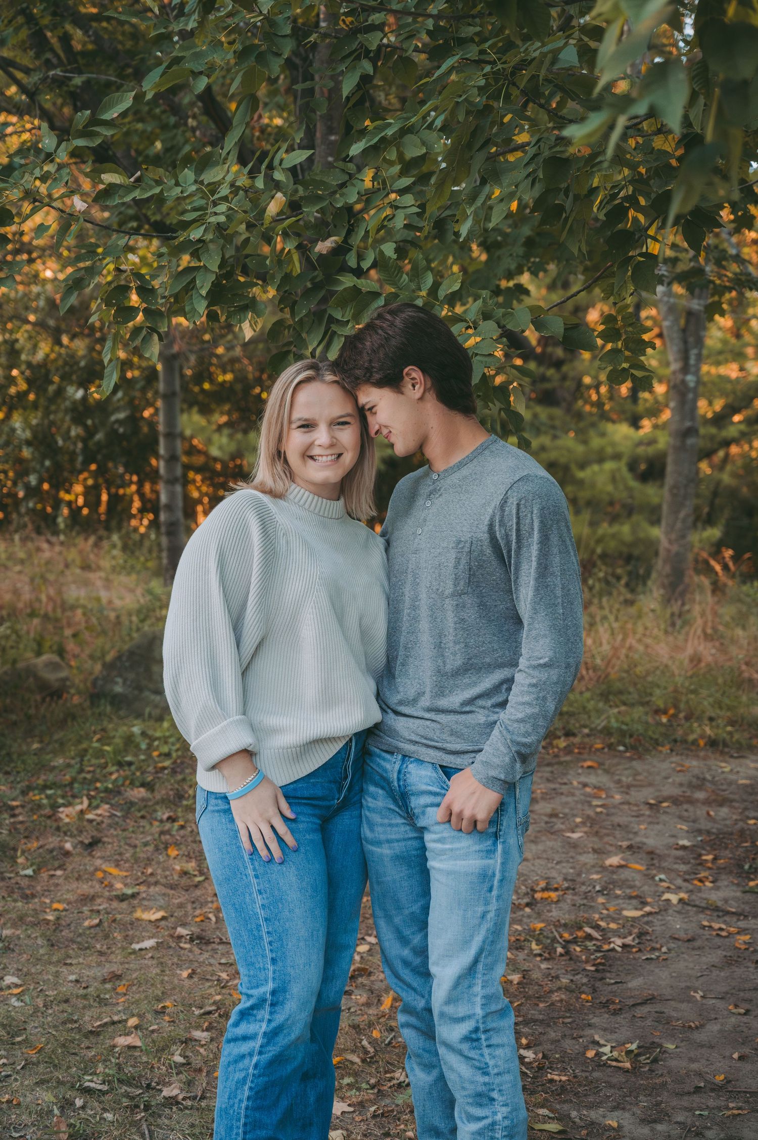 A couple shares a tender moment among fall trees wearing matching sweaters and jeans.