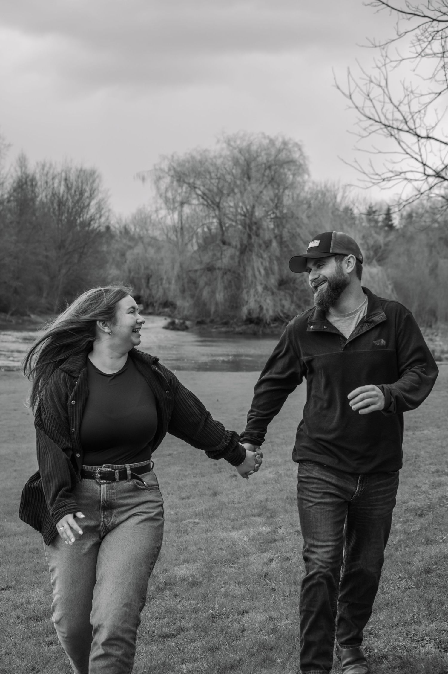 Black and white photo of a smiling couple holding hands and walking outdoors near a river with trees in the background.