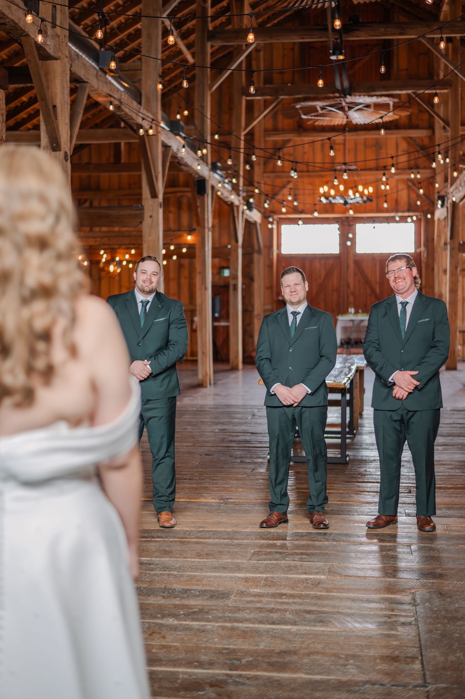 Three groomsmen in matching green suits stand in the rustic Brussels Four Winds Barn decorated with string lights.