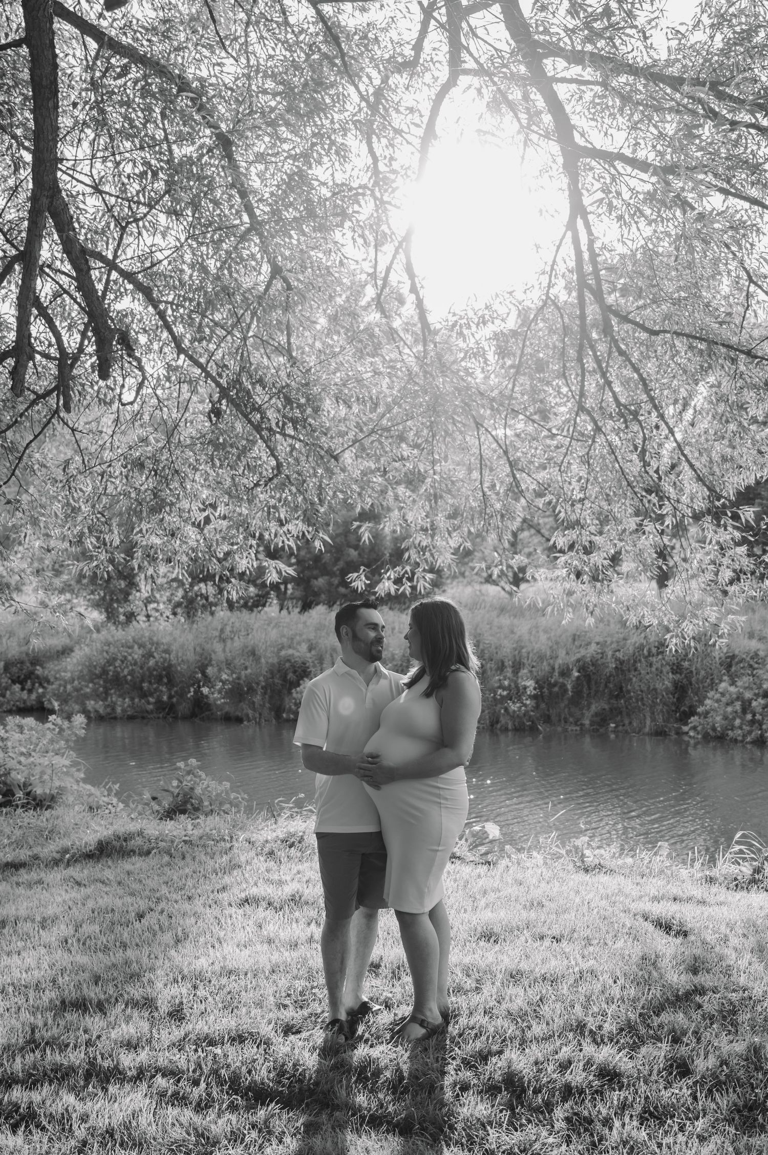 A black and white sequence shows a couple embracing outdoors under flowering trees by a tranquil pond during their maternity session.