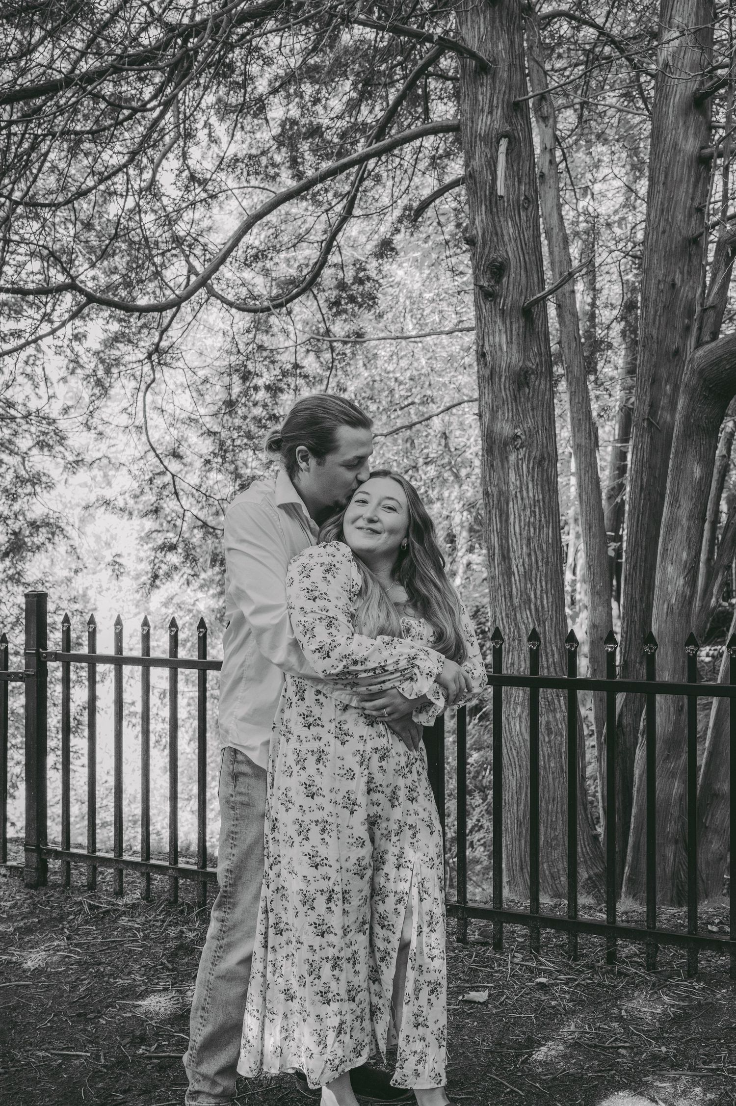 A tender black and white moment between a couple in a wooded setting with iron fencing.