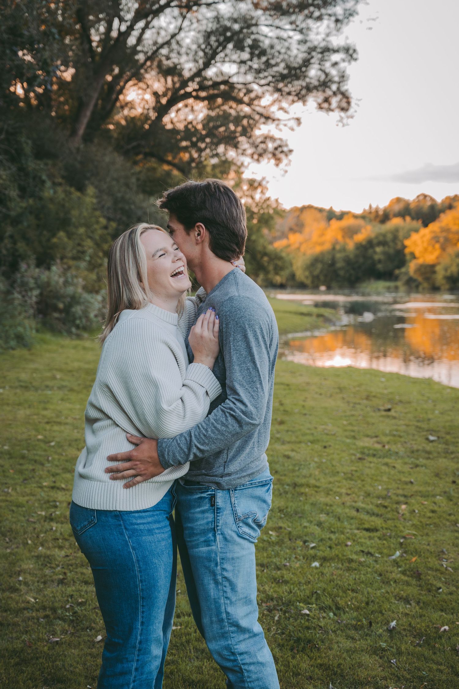 A couple embracing at sunset by a lake with autumn trees creating a romantic backdrop.