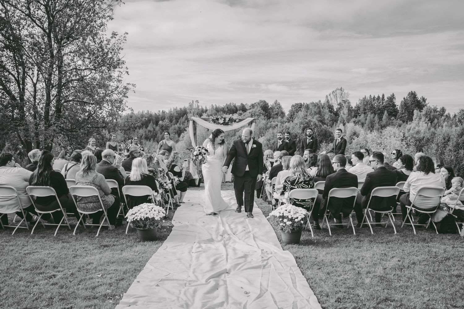 Black and white photo of newlyweds walking down aisle with guests seated on both sides at outdoor ceremony.