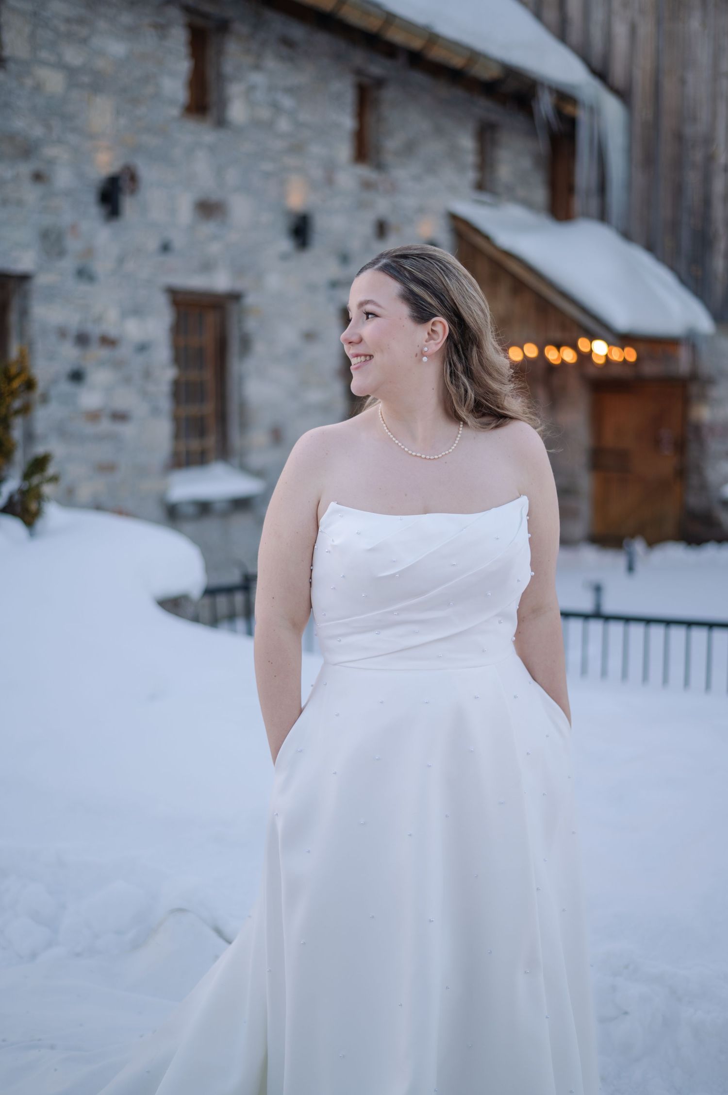 Bride in strapless white gown smiling outdoors in winter snow, rustic stone building with warm lights behind her.