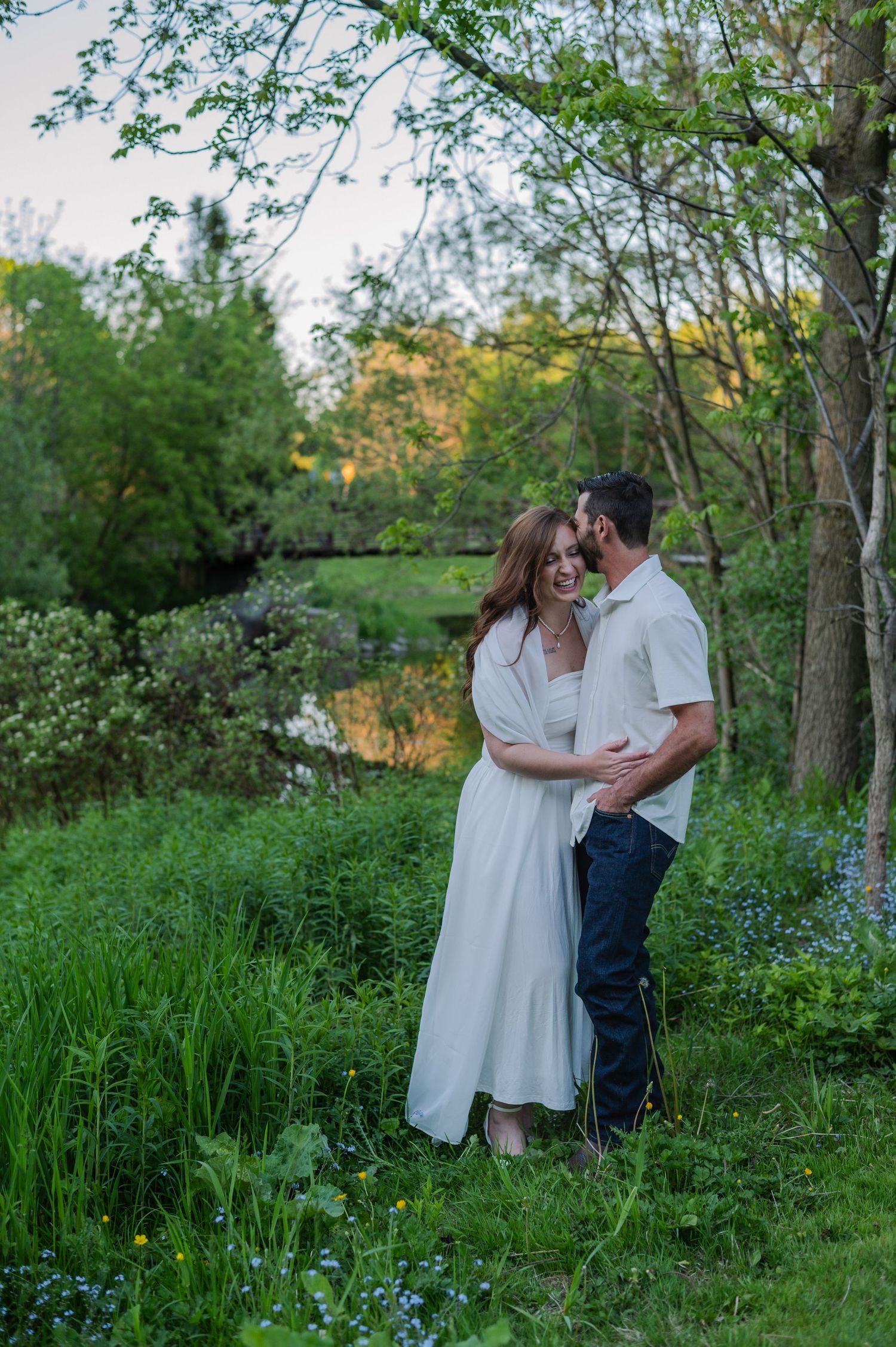 Couple embracing in lush green garden near pond at sunset, woman in white dress, man in jeans and white shirt.