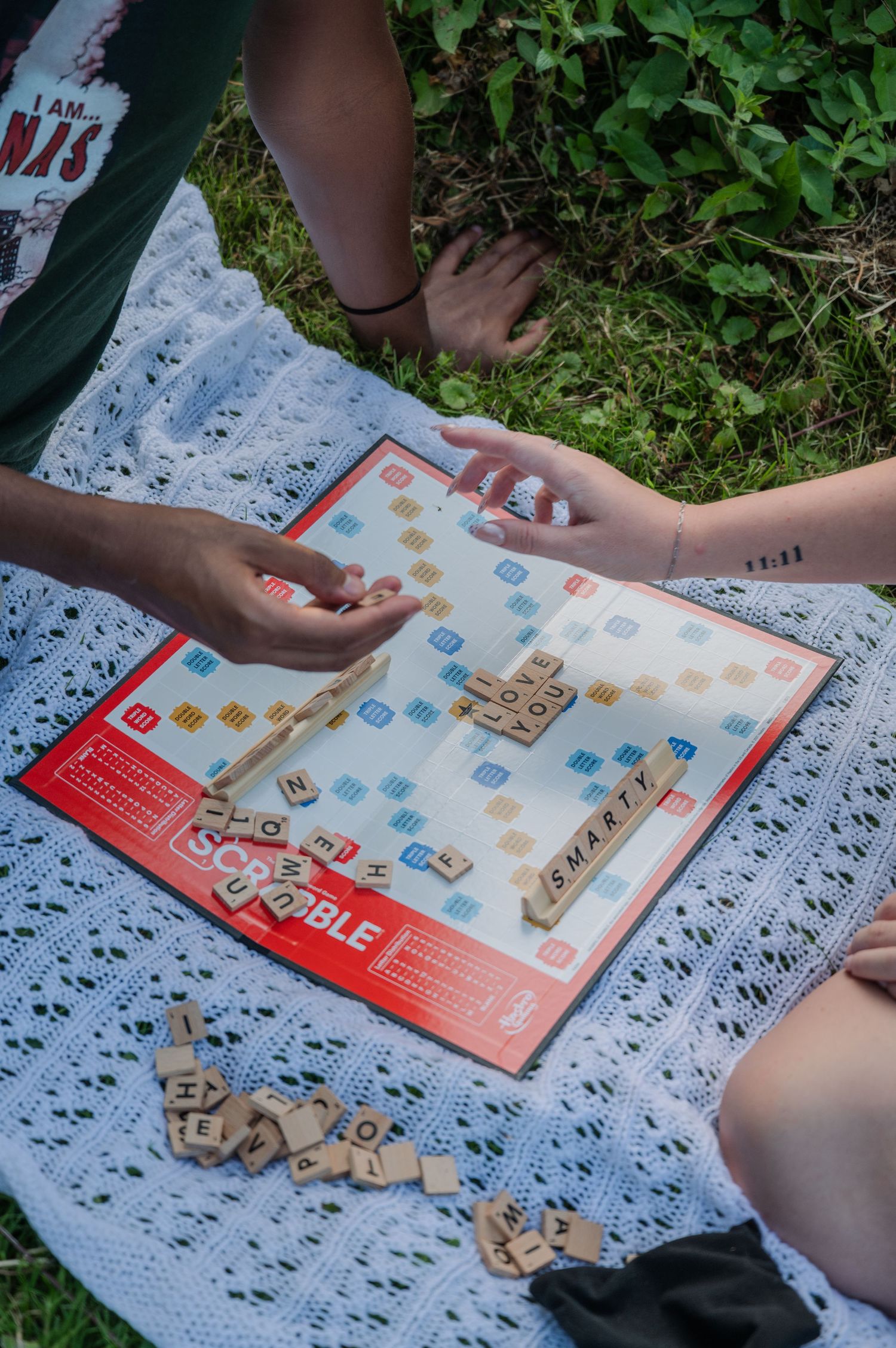 Two people playing Scrabble outdoors on a white blanket in the grass, with tiles spelling SMART visible on the board.
