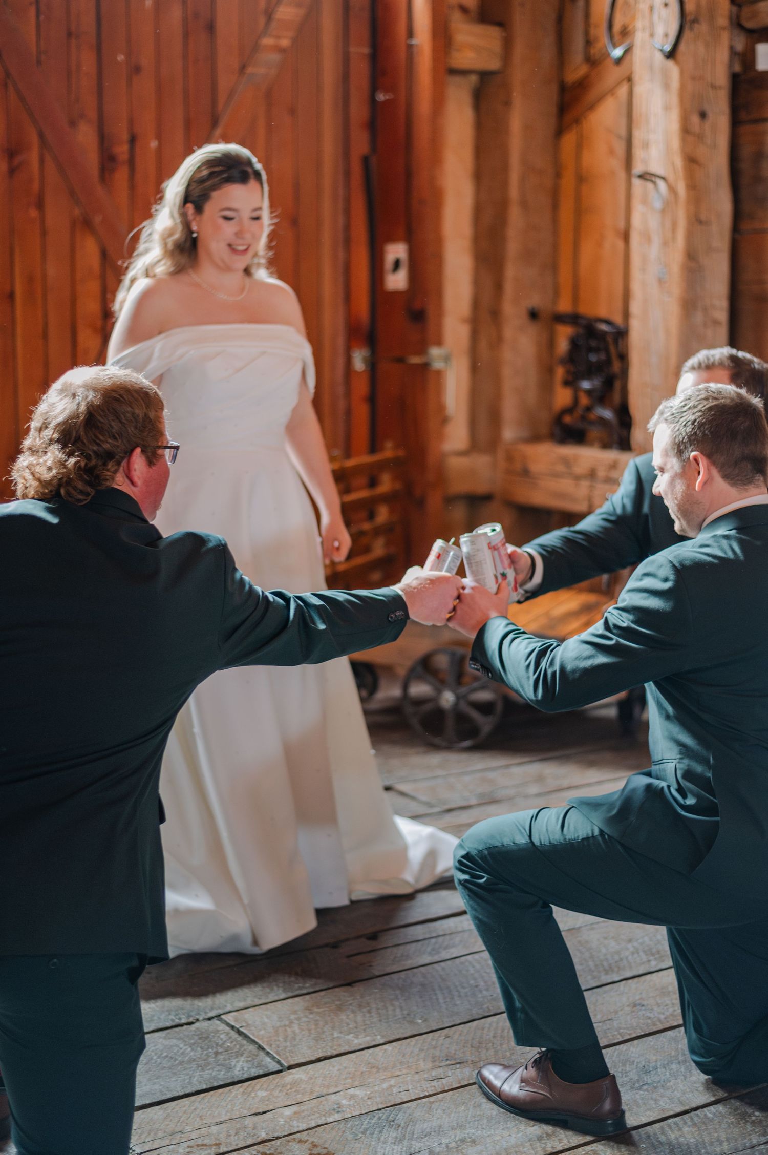 Two groomsmen kneel before a bride in a white off-shoulder gown inside a rustic barn wedding venue.