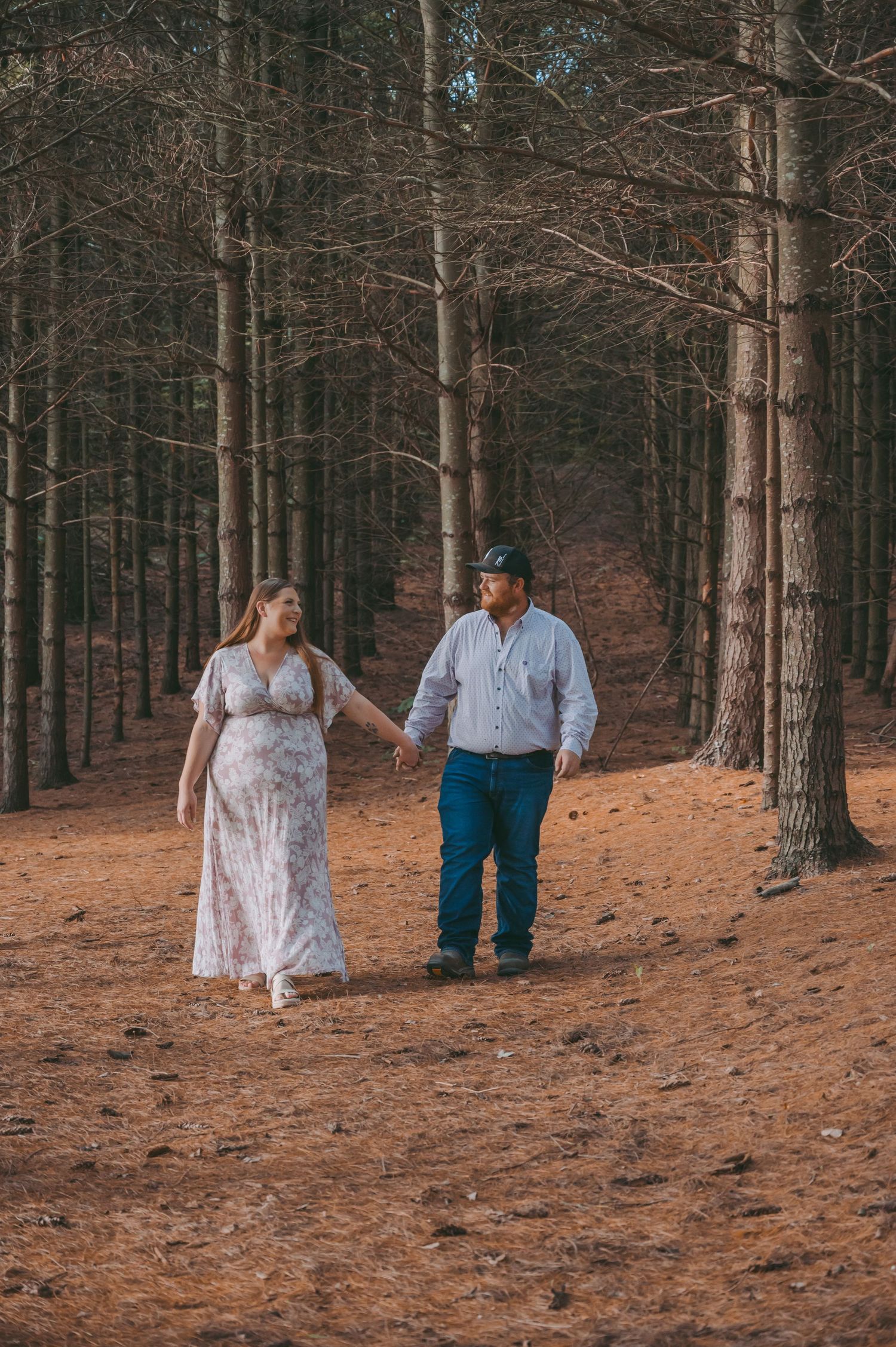A couple walks hand in hand through a pine forest on a dirt path.