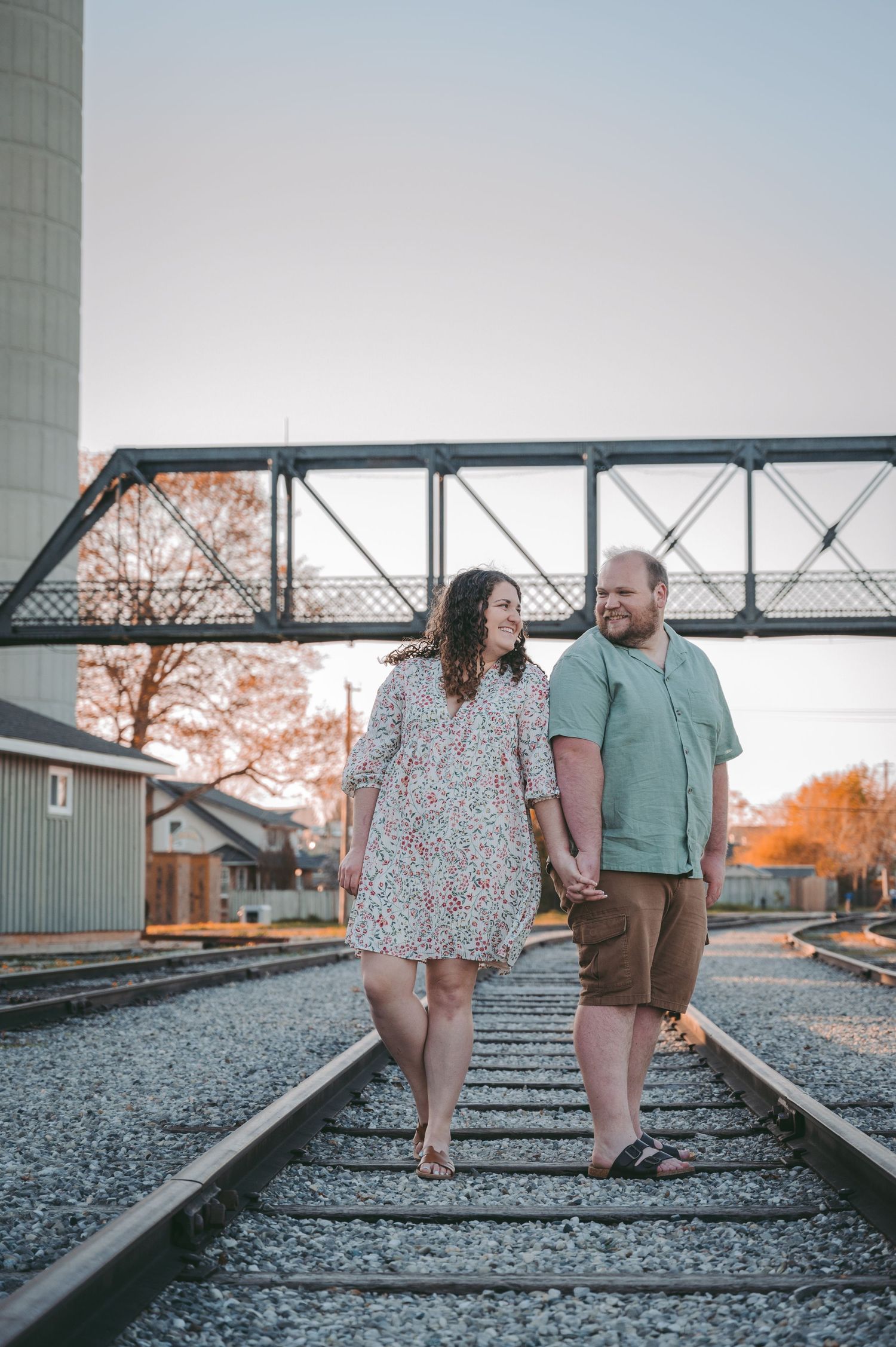 A couple stands together on railroad tracks with an industrial backdrop at sunset.