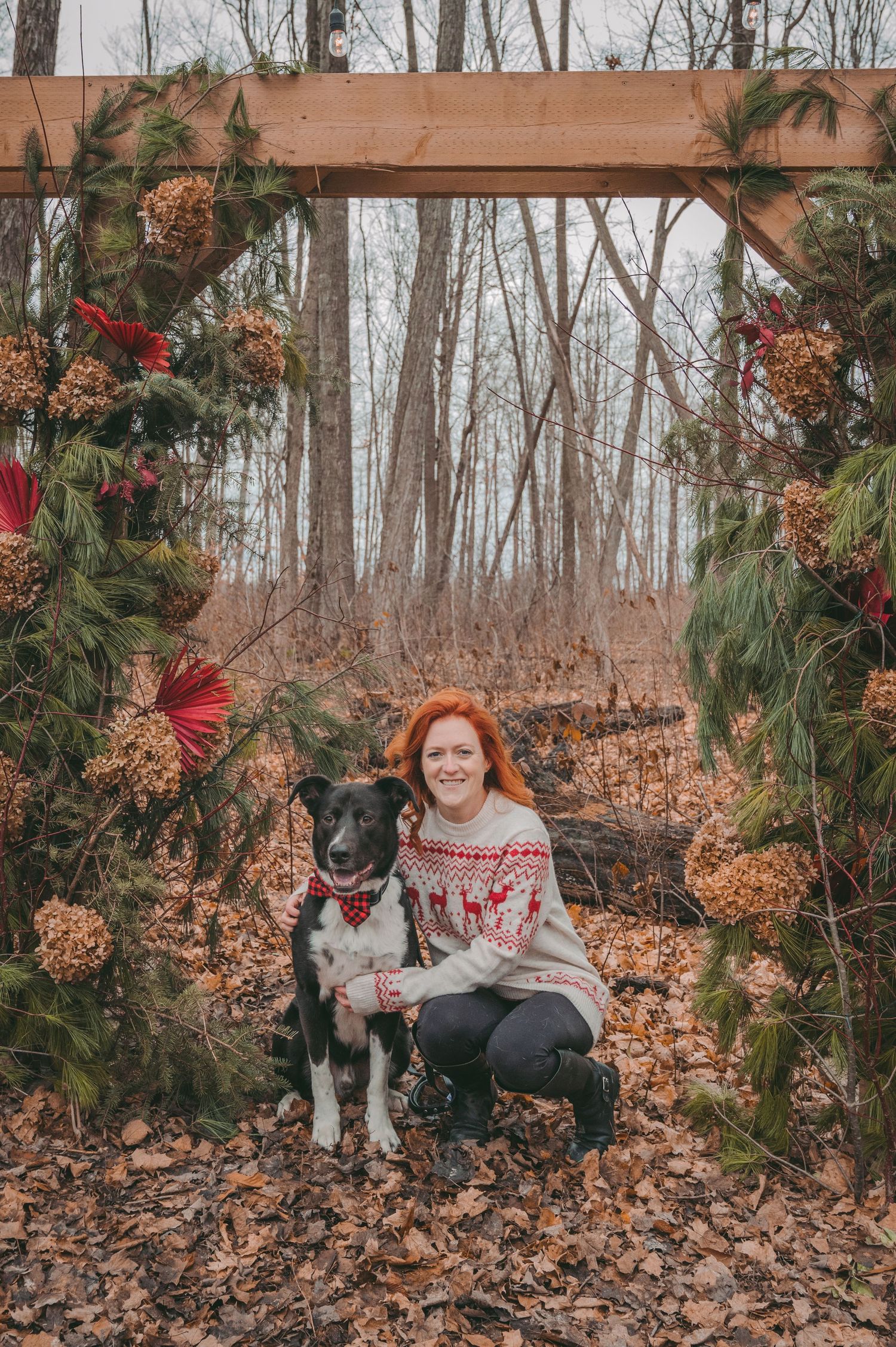 Winter portrait featuring person in holiday sweater with black and white dog under decorated wooden arch.
