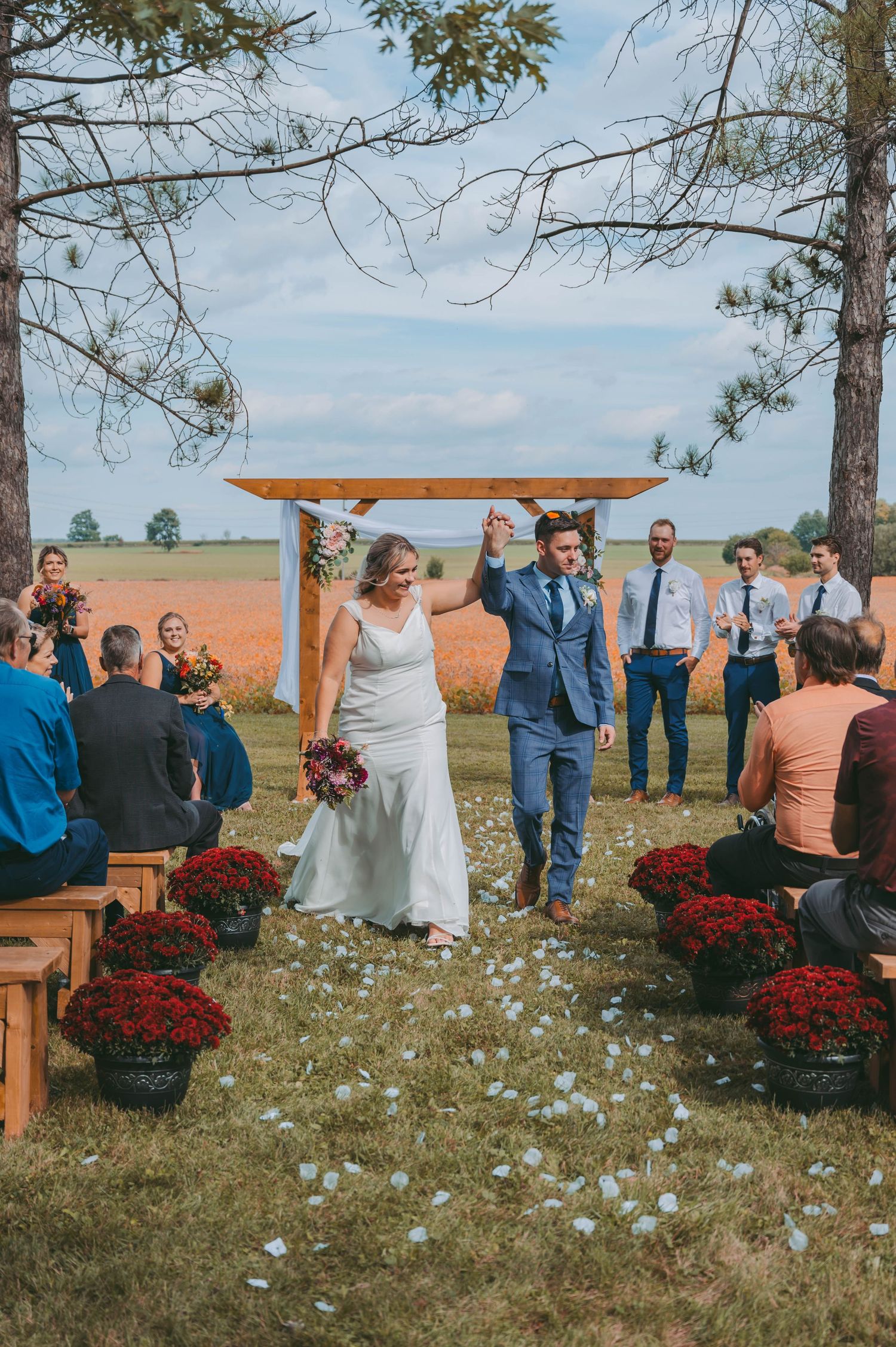 Outdoor wedding ceremony setup with wooden benches and red mums in a field under pine trees.