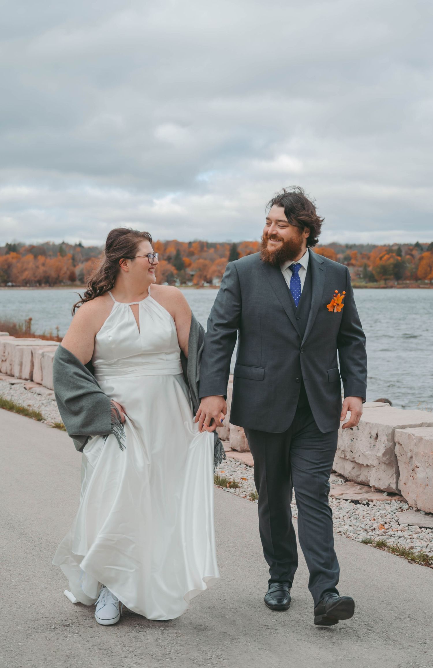 Newlyweds walking along lakeside path with autumn colors reflecting on water.
