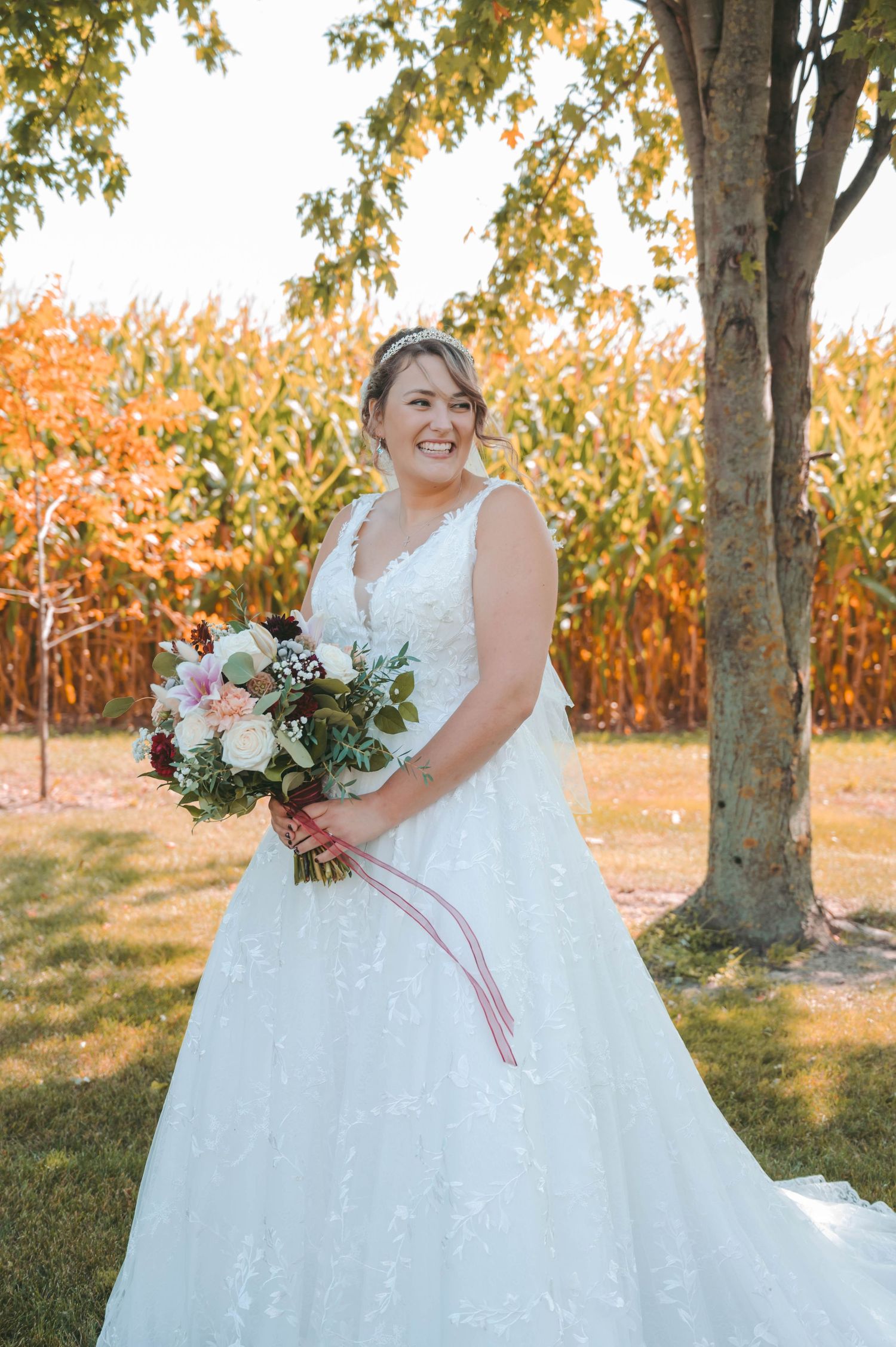 Bride in lace white wedding dress holds floral bouquet while standing next to autumn cornfield and tree.