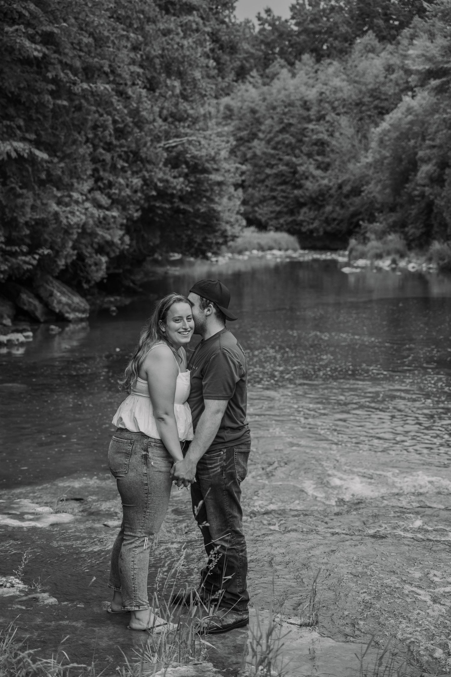 A romantic moment captured in black and white as a couple embraces by a serene river surrounded by trees.