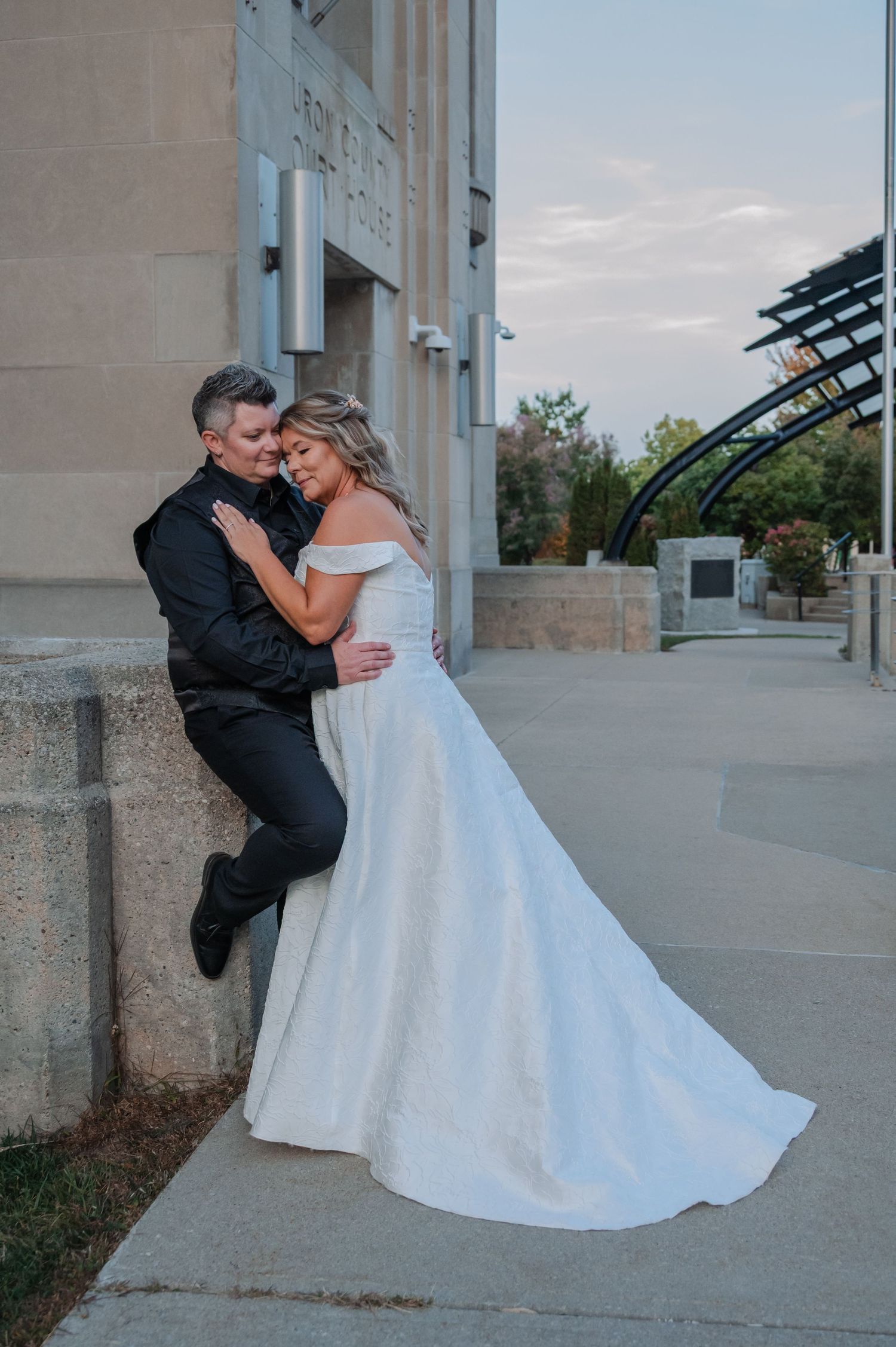 Bride in off-shoulder white gown embraces partner in black suit against stone building at dusk.
