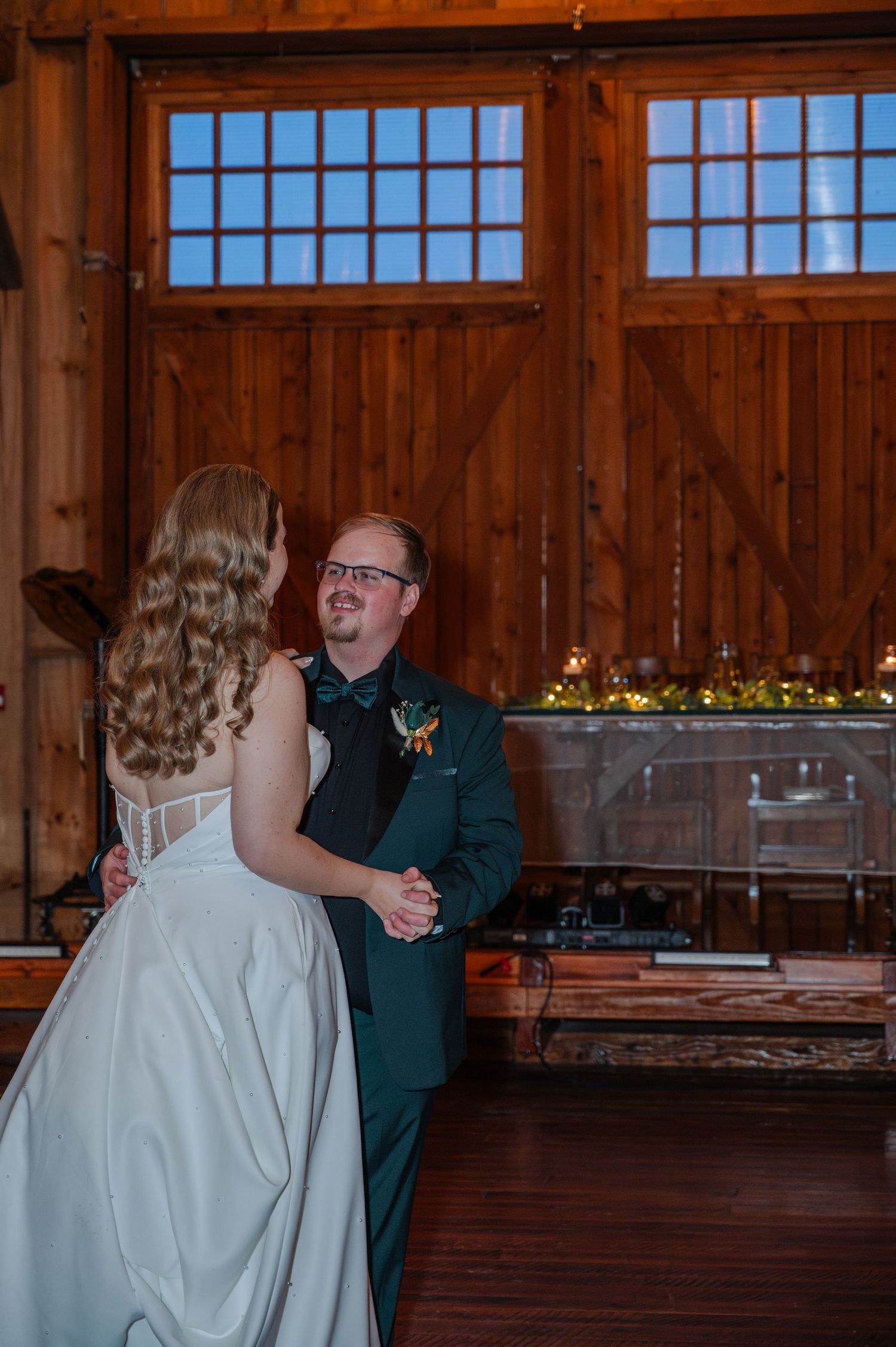 Bride and groom share their first dance in a rustic barn venue, with wooden walls and large windows in the background.
