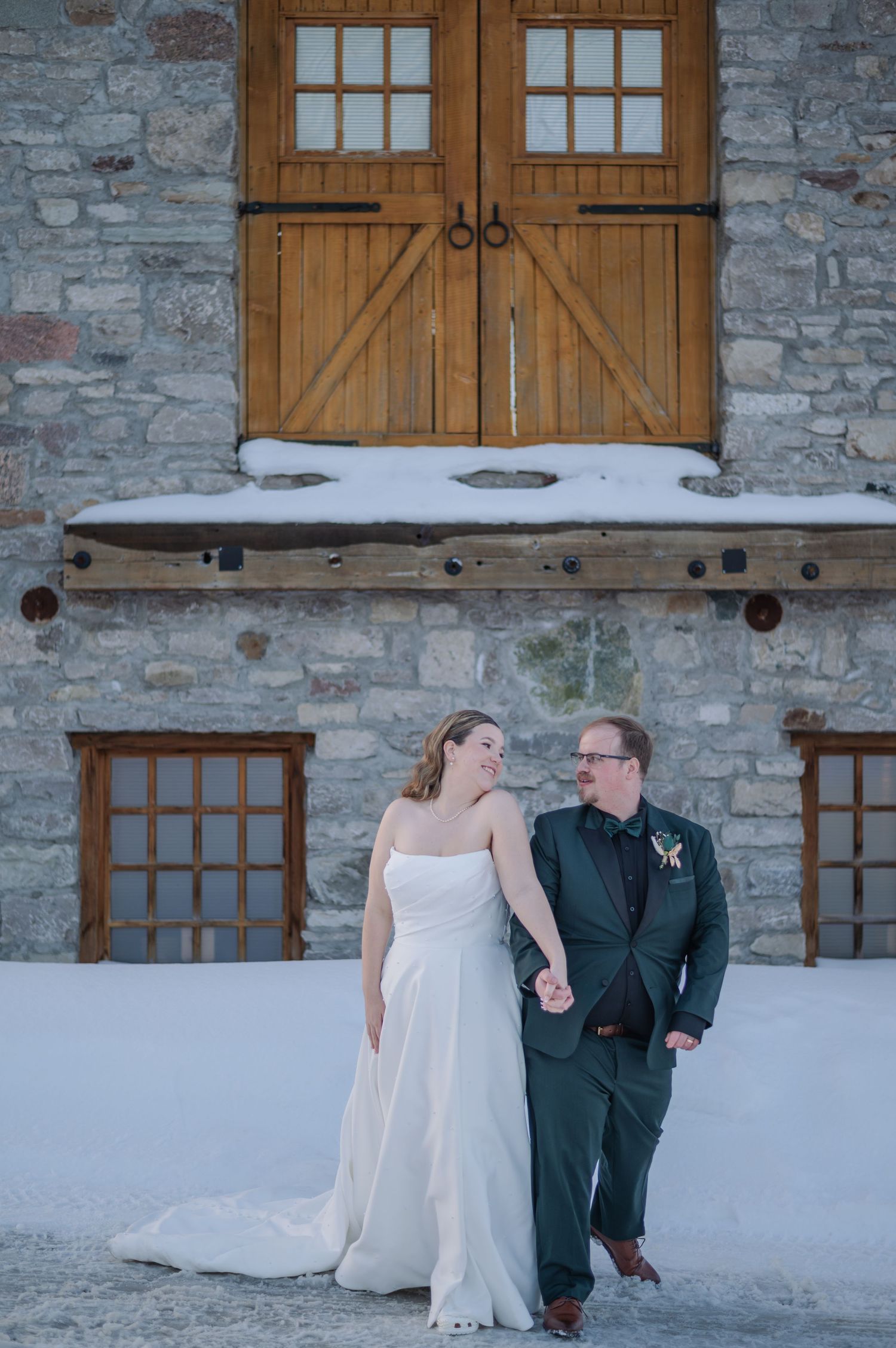 A couple together against a rustic stone wall with wooden shuttered windows.