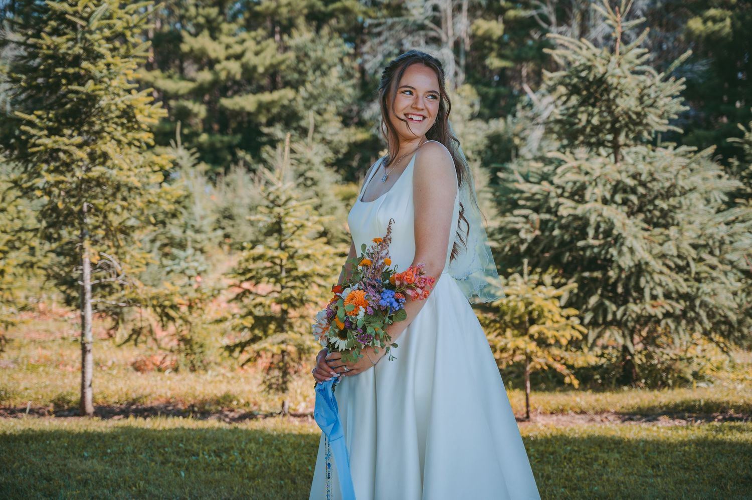 Bride in white dress holding colorful bouquet against forest backdrop.