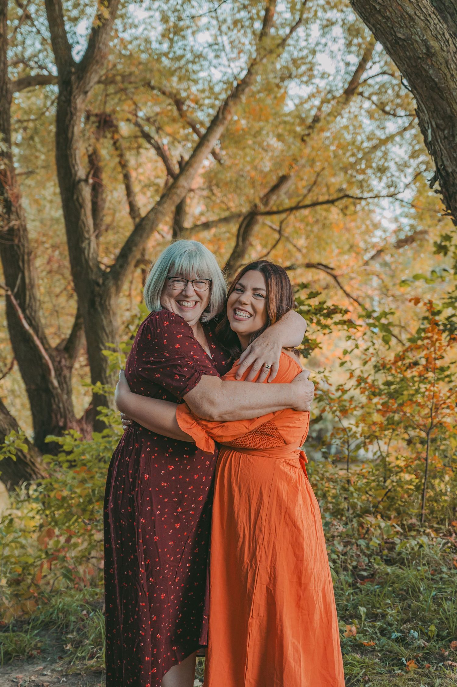 Women in orange and burgundy dresses share joyful hugs in an autumn woodland setting.